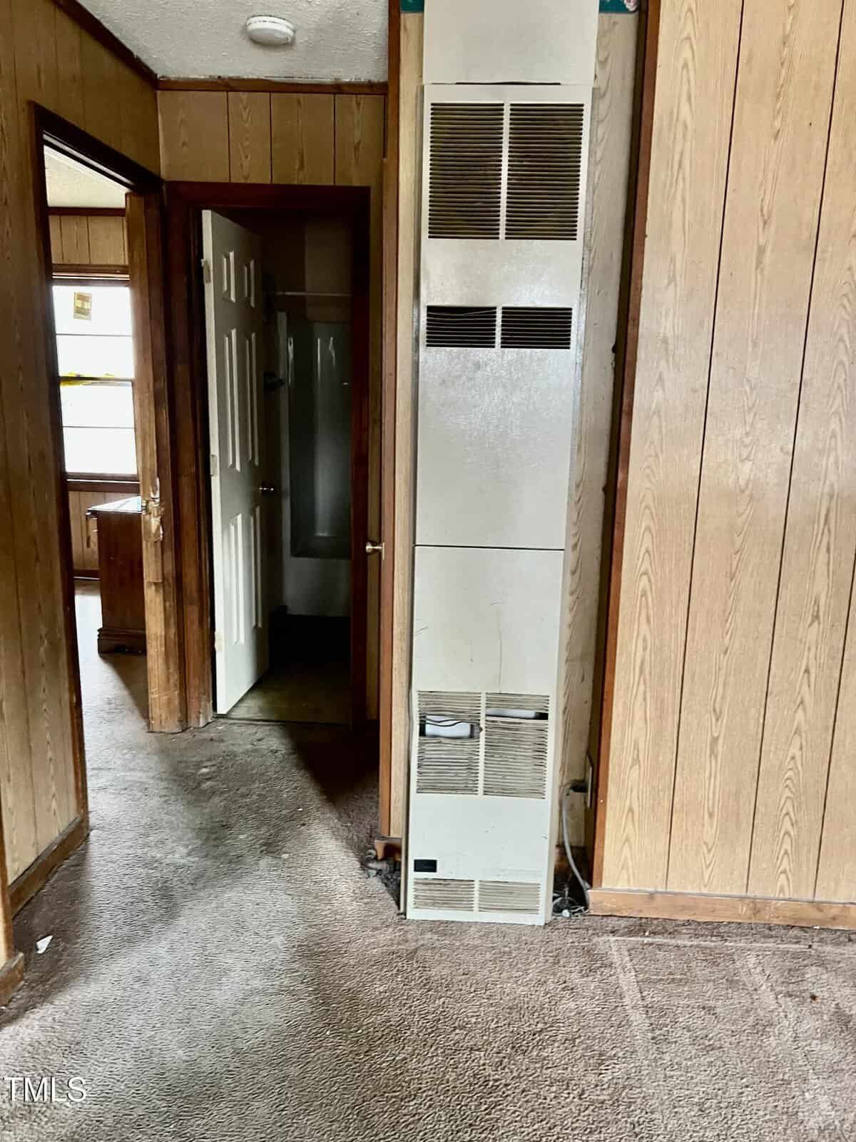 A narrow, wood-paneled hallway features a tall, white wall heater in the center. Open doors on either side lead to other rooms. The carpeted floor appears worn and the walls show signs of age.