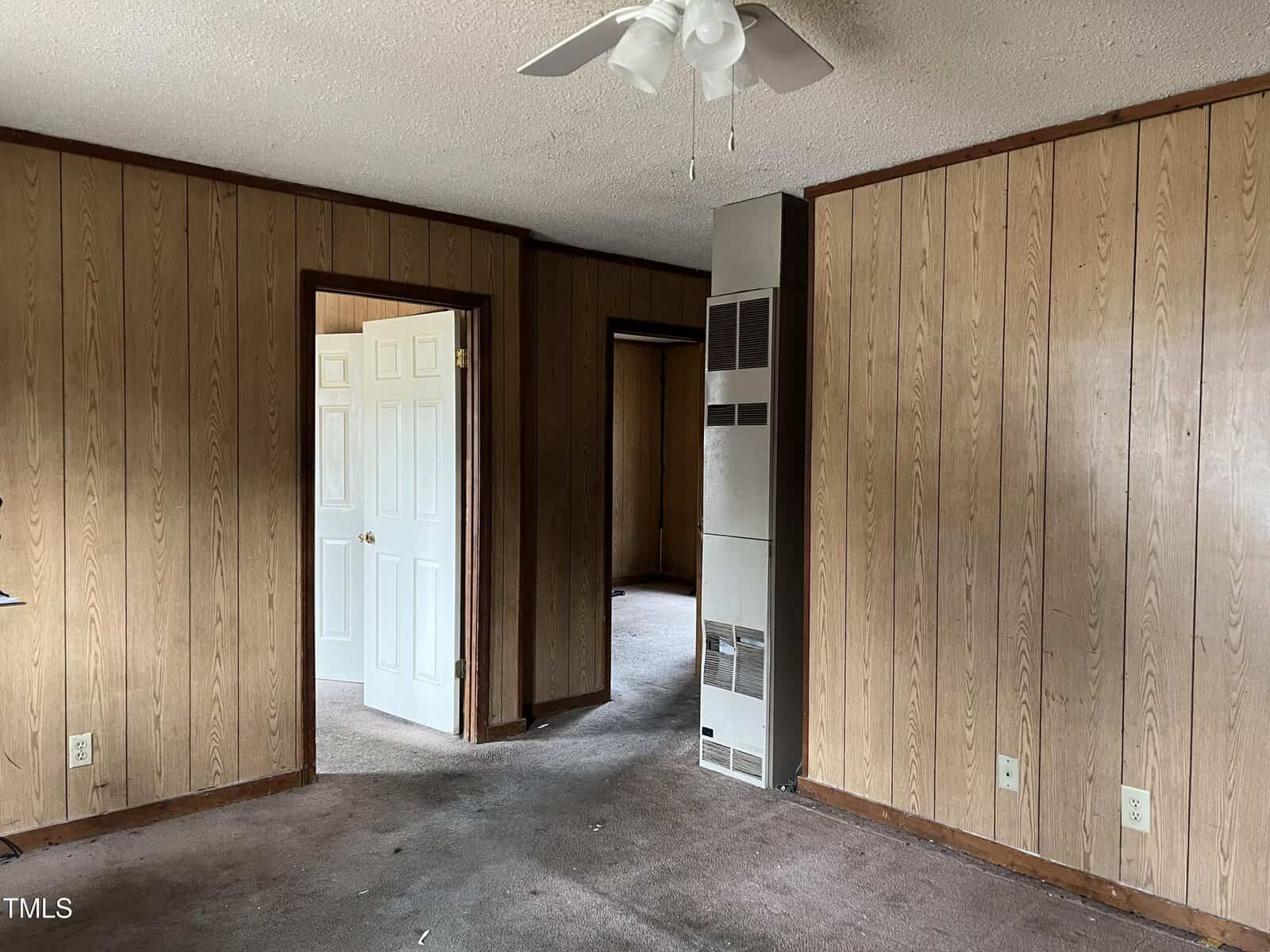 A room with wood-paneled walls and a ceiling fan. There is a doorway leading to another room with an open white door. The carpet is worn and the room appears empty. A vertical metal heater is visible in one corner.