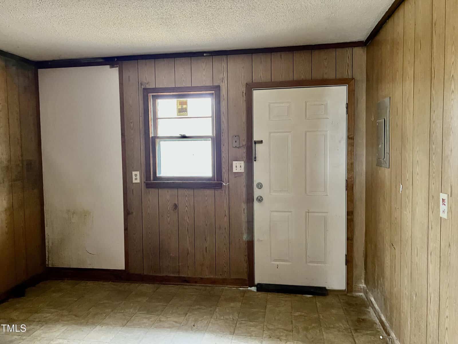 Room with wood-paneled walls and a textured ceiling. A window with blinds is on the left, above a small section of white wall. A closed white door with a mail slot is next to the window. The floor has a tan linoleum covering.