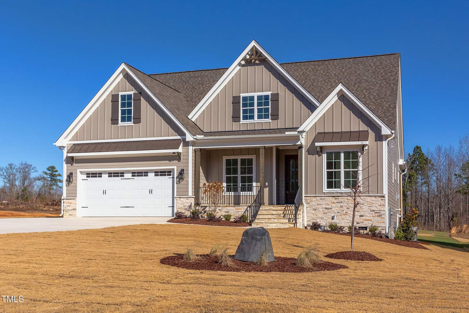 A modern two-story house with tan siding, white trim, a covered front porch, and an attached two-car garage, surrounded by a neatly landscaped yard and driveway under a clear blue sky.