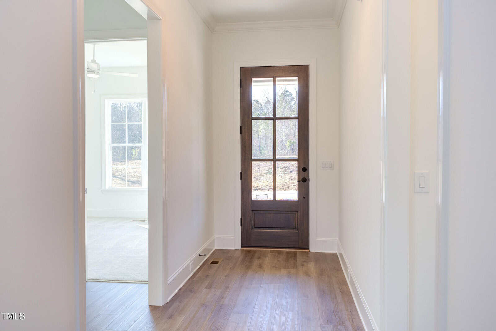 A hallway with light wood flooring leads to a wooden front door with glass panels. To the left, an open doorway reveals a bright room with a large window and a ceiling fan. Walls and trim are painted white.