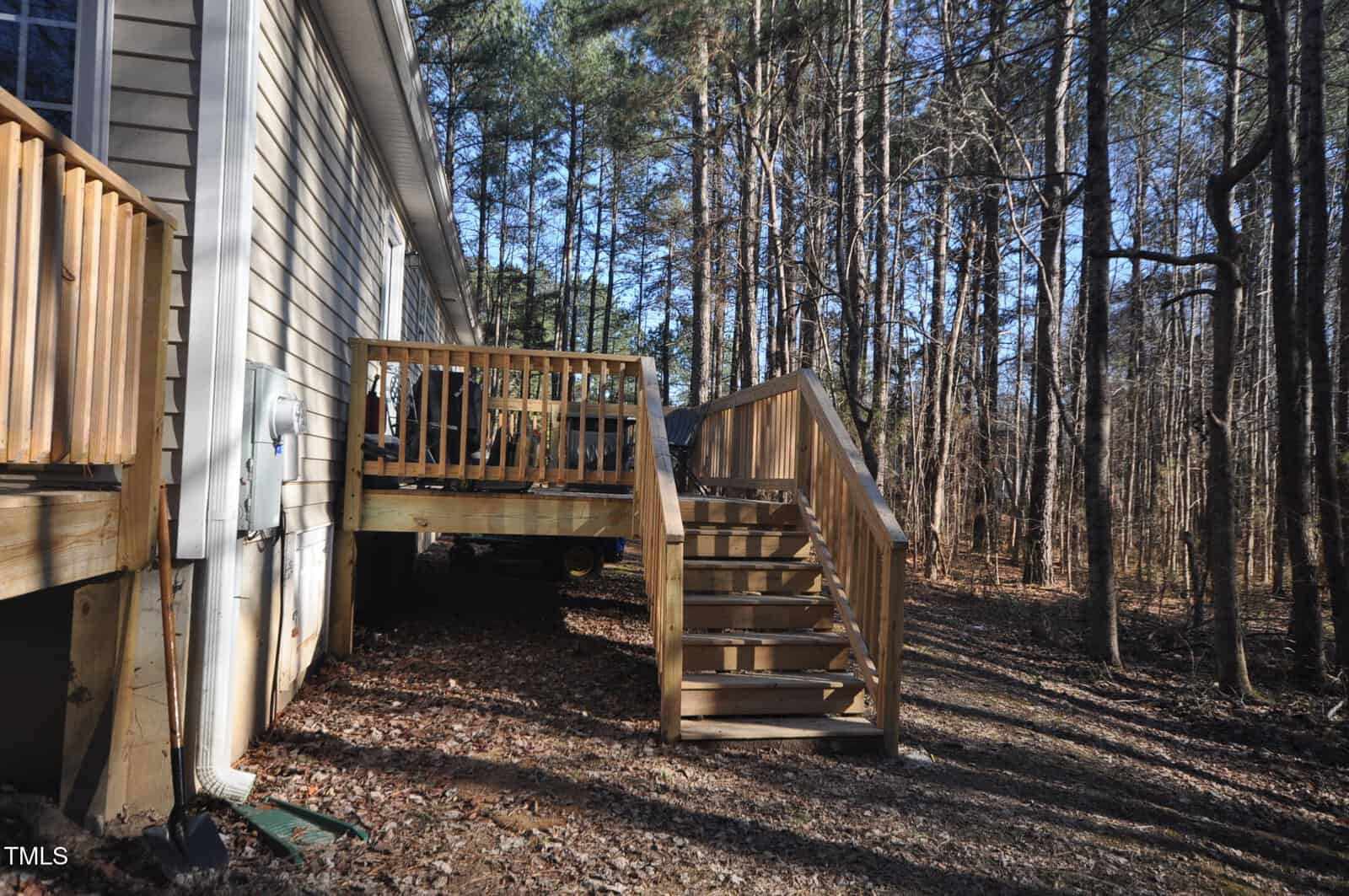 A wooden deck with a staircase leads to the side of a house. The house is in a wooded area, surrounded by leafless trees. The ground is covered with brown leaves and some patches of grass. The sky is clear, and sunlight filters through the trees.