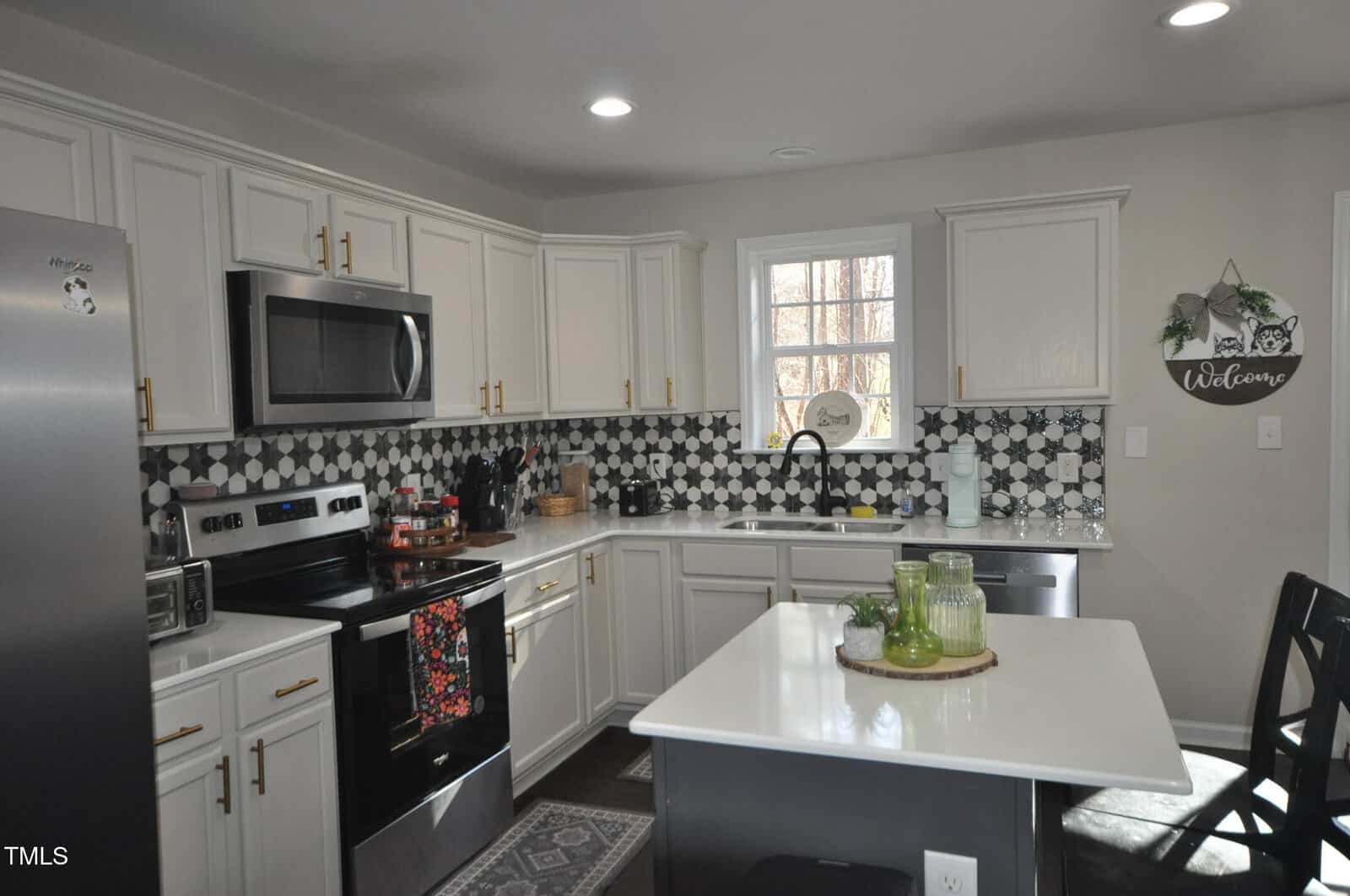 Modern kitchen with white cabinets and a black and white geometric tile backsplash. Stainless steel appliances, including a refrigerator and oven, are present. A white island with green decor items and a cushioned chair is in the center.