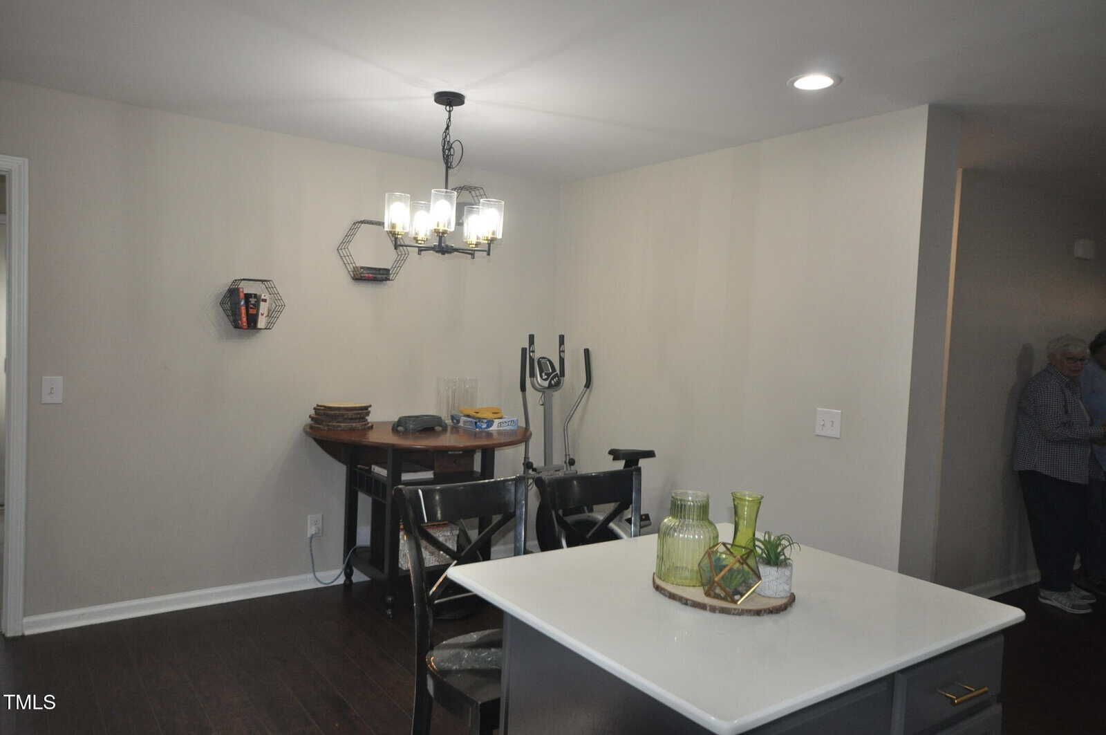 A modern dining area features a table with two chairs, a hanging light fixture, and an exercise bike against the wall. There are decorative items on a small wall shelf and a kitchen island in the foreground. A person is partially visible to the right.