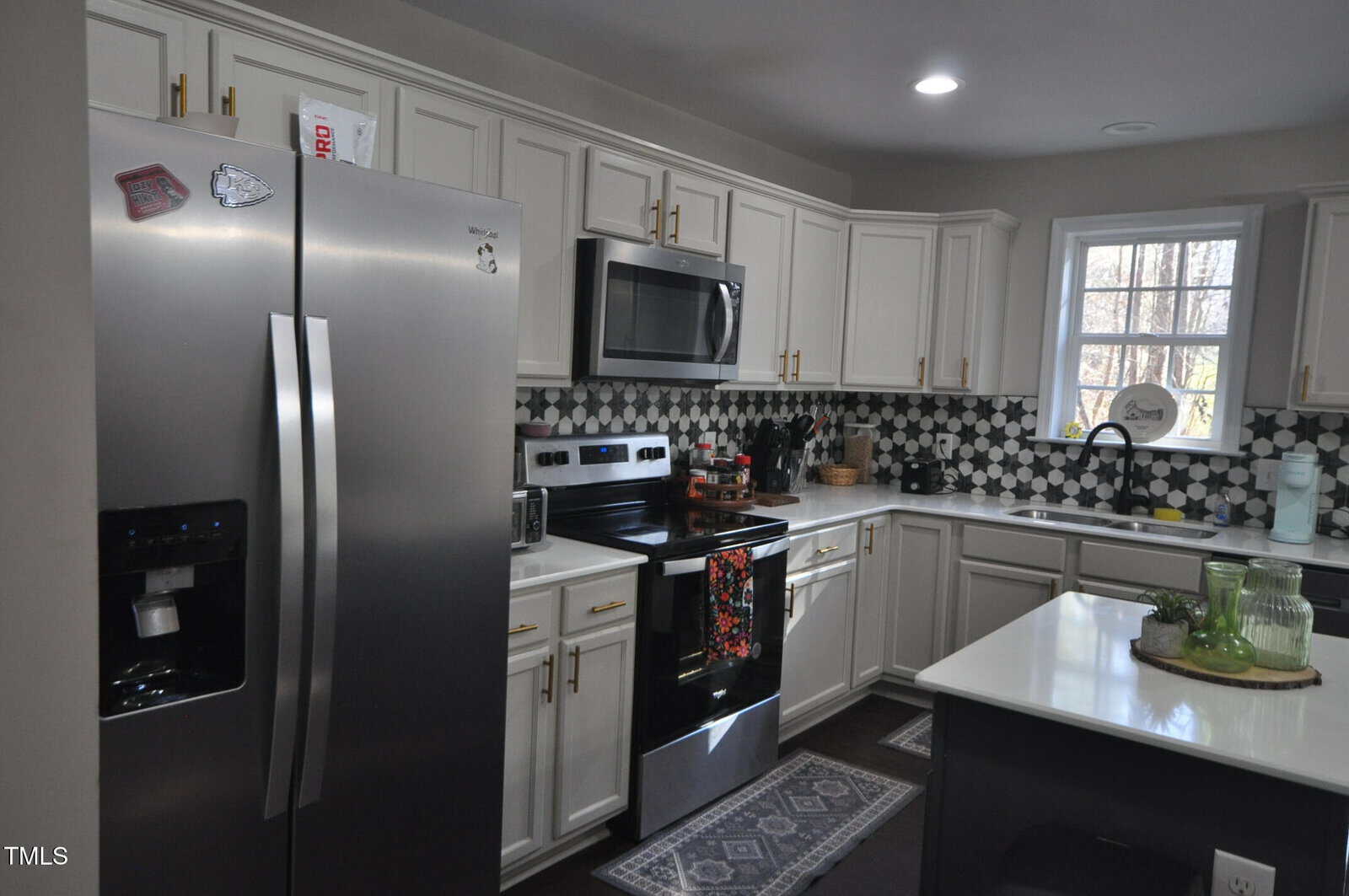 Modern kitchen with white cabinets and dark countertops. Stainless steel refrigerator and oven are present. The backsplash features a geometric pattern. A small island holds green glassware, and a window allows in natural light.
