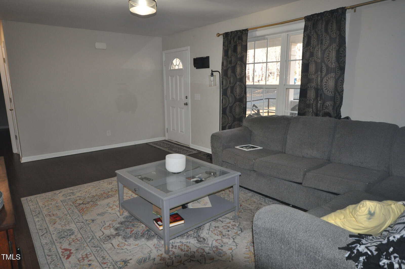 Living room with gray walls and dark wood floor. A gray sectional sofa and coffee table are placed on a patterned area rug. A large window with dark curtains faces the sofa. A small ceiling light hangs near the entrance door.