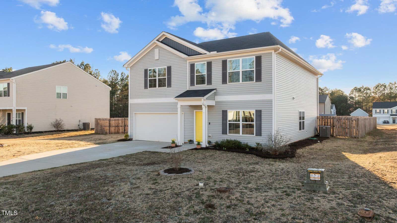 A two-story light gray house with white trim and a yellow front door, featuring a driveway, small front yard, and a fenced backyard, under a blue sky with scattered clouds.