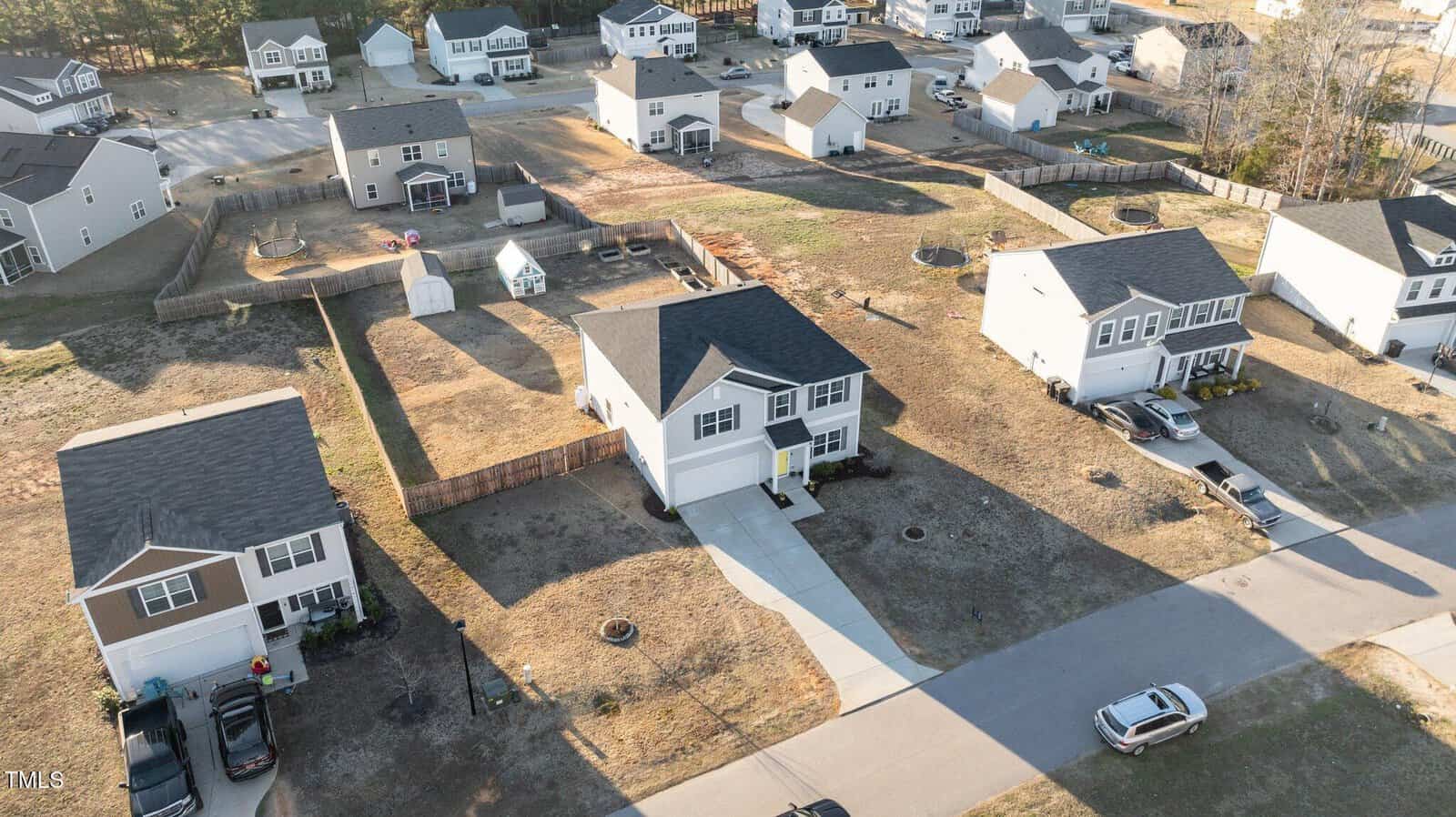 Aerial view of a suburban neighborhood with several two-story houses, driveways, parked cars, fenced backyards, and a small playground area. The lawns are dry and there are few trees.