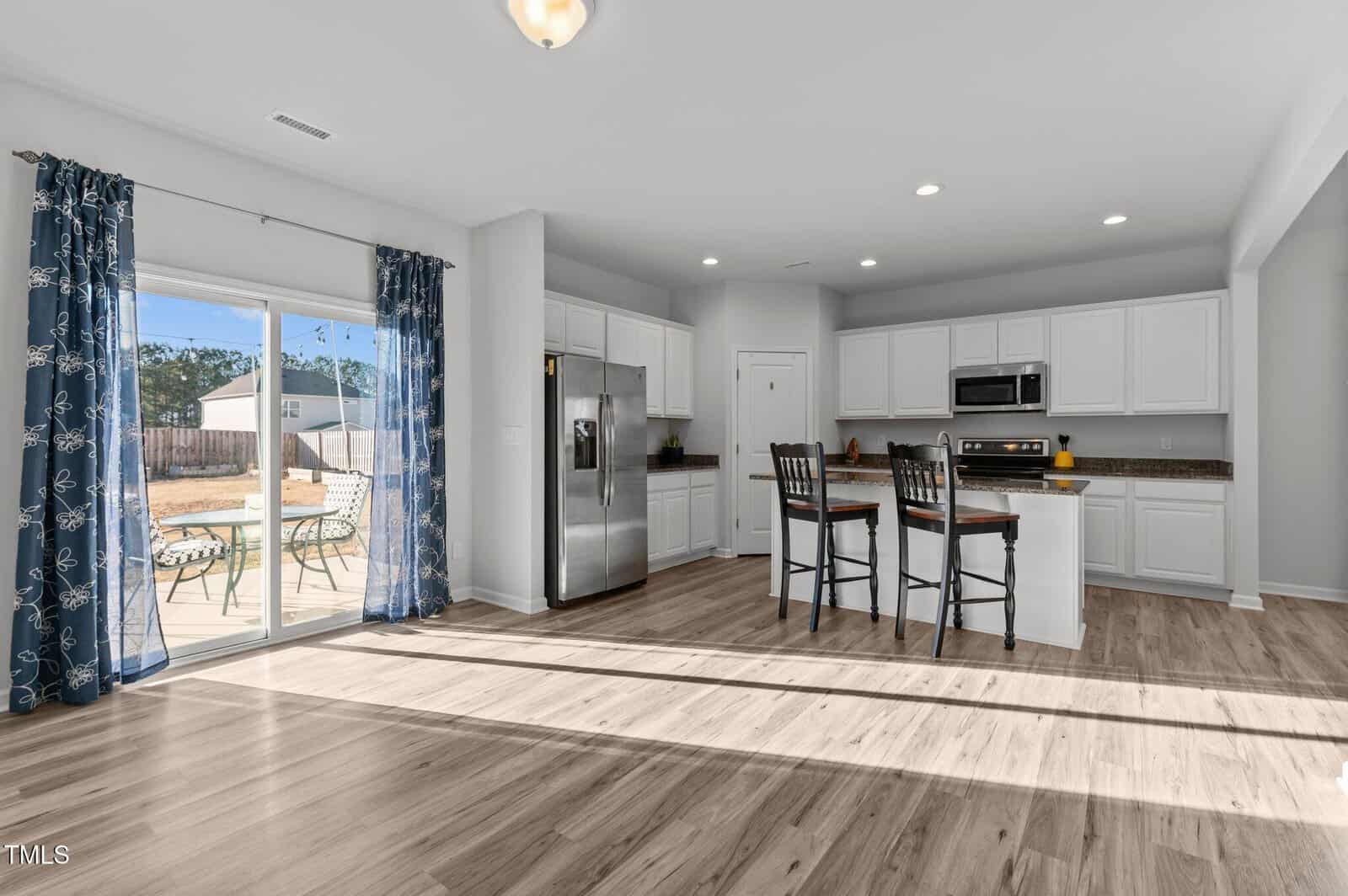 Modern kitchen with white cabinets, stainless steel appliances, island with two chairs, and brown countertops. Large glass doors with blue curtains open to a patio and backyard. Light wood floors throughout the room.