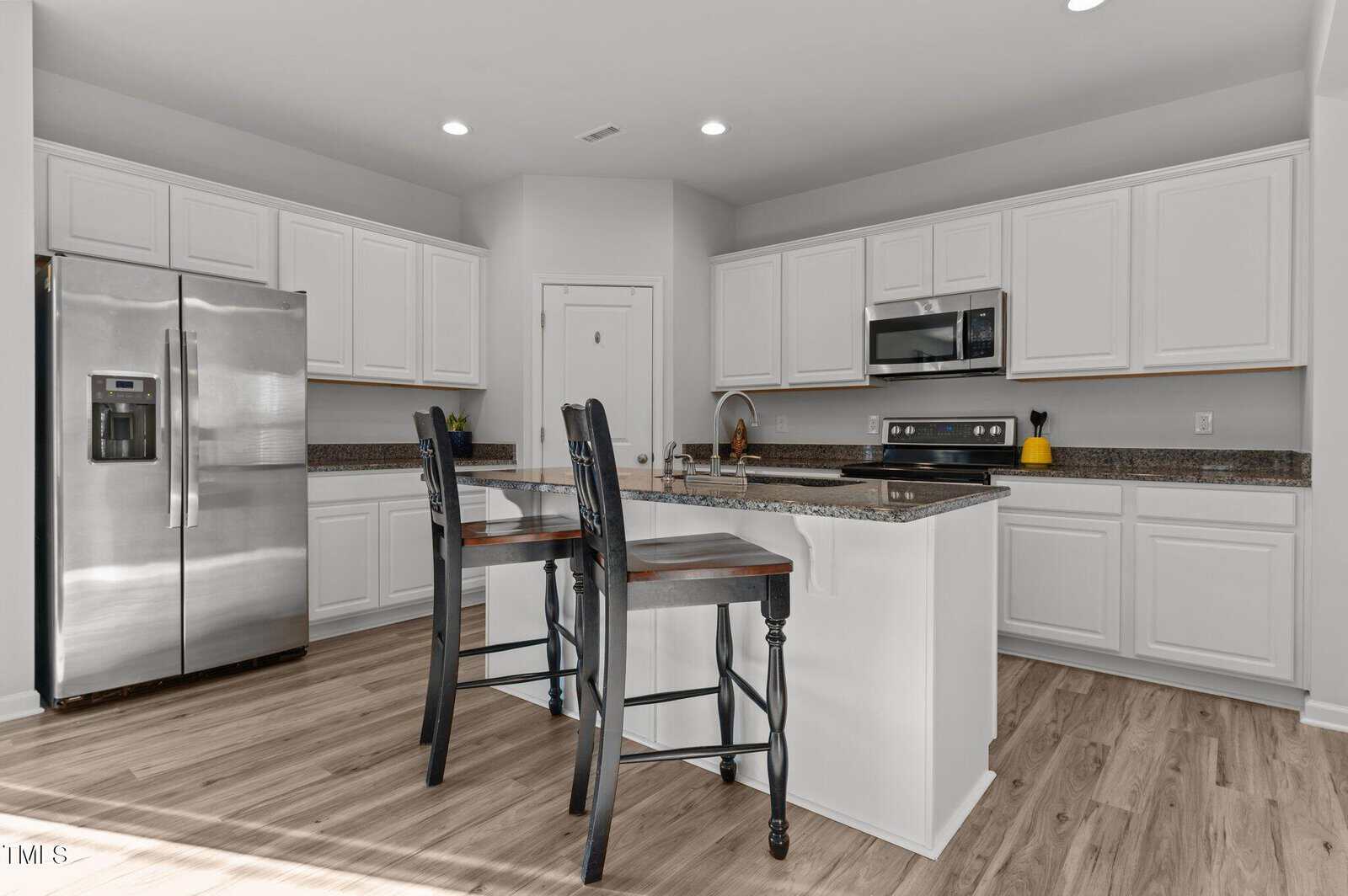 Modern kitchen with white cabinets, stainless steel appliances, granite countertops, an island with a sink, and two black bar stools. Light wood flooring and recessed lighting are also visible.