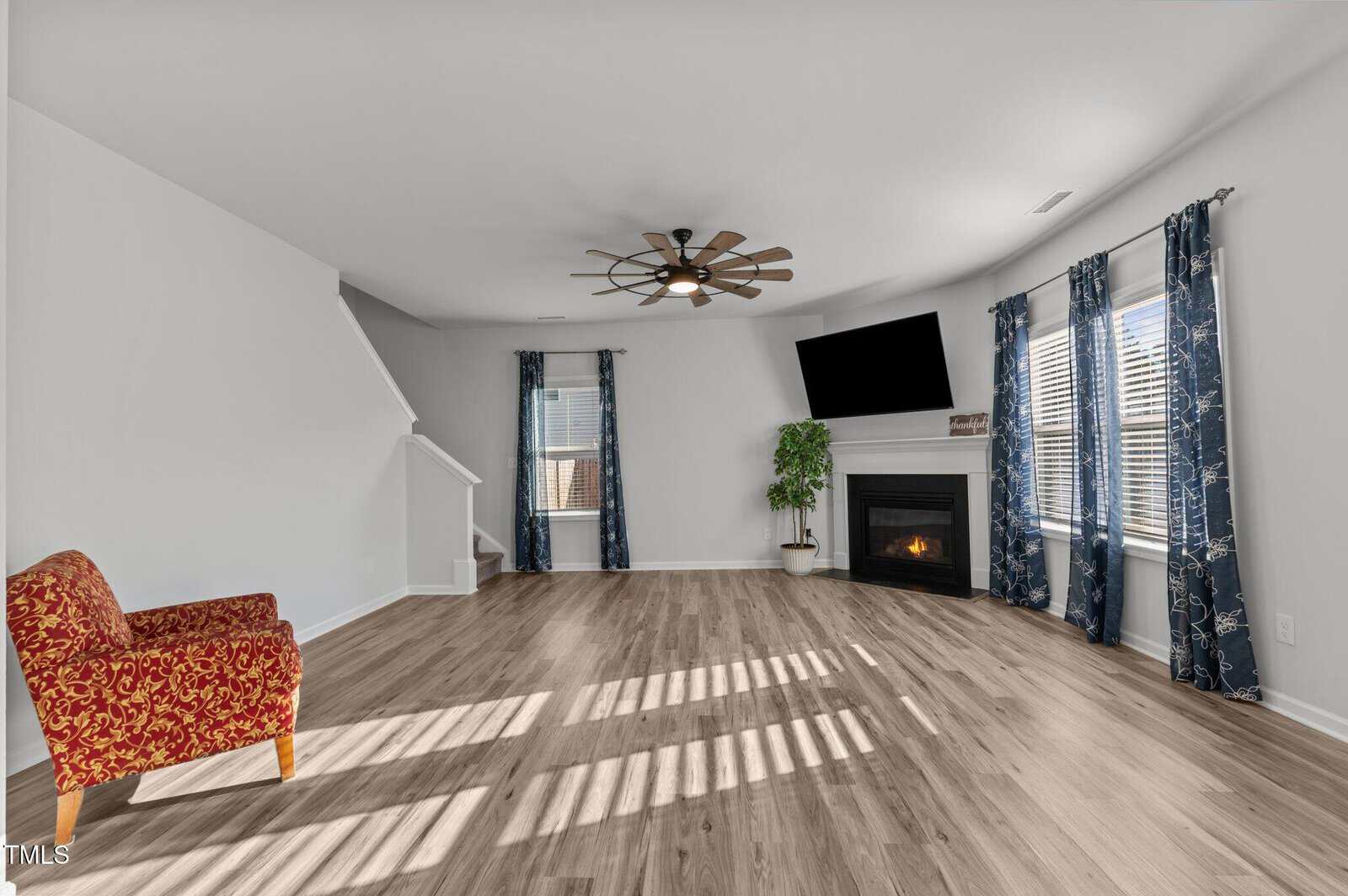 A spacious living room with light wood floors, a red patterned armchair, a corner fireplace with a TV above it, blue curtains on windows, a green plant, and a modern ceiling fan. A staircase is visible in the back.