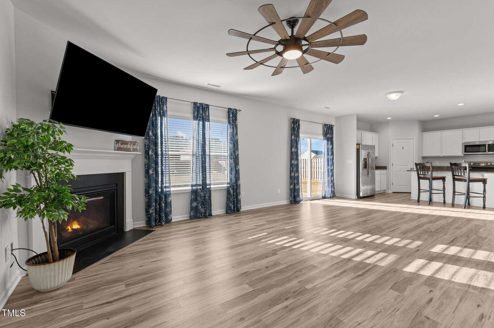 Bright, open living area with light wood floors, a fireplace, large wall-mounted TV, windows with blue curtains, a ceiling fan, a potted plant, and a kitchen with white cabinets and an island with barstools.