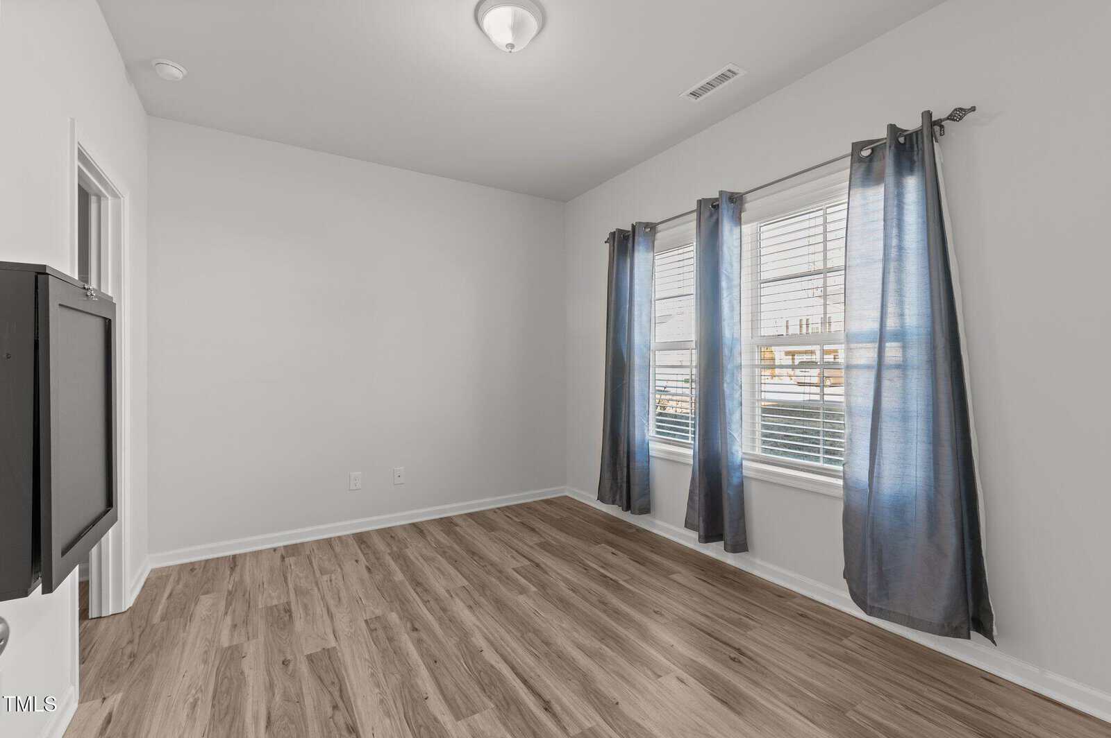 An empty room with light gray walls, wood-style flooring, a ceiling light fixture, and two windows with gray curtains letting in natural light. A wall-mounted unit is visible on the left wall.