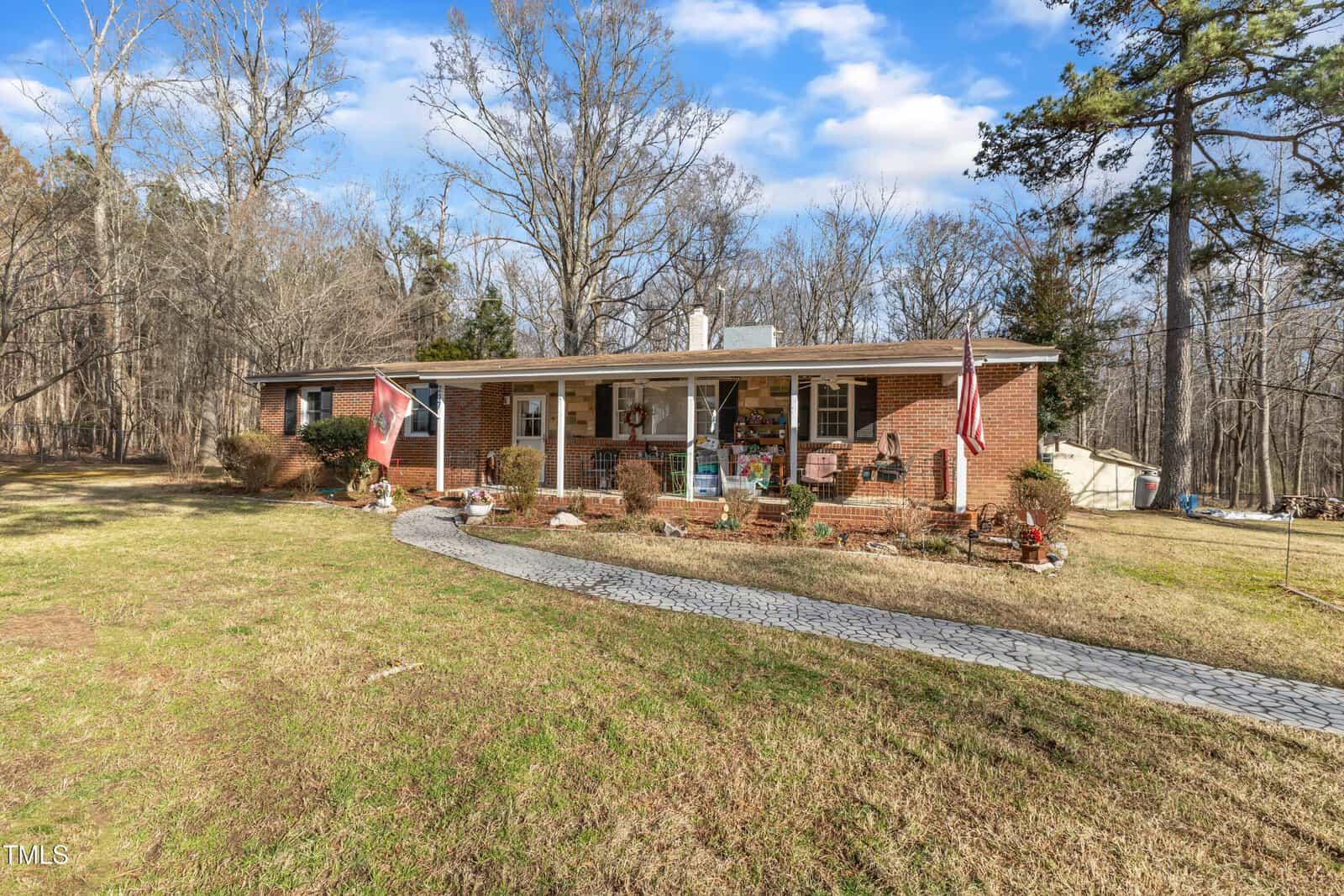 A single-story brick house with a covered front porch, decorated with flags and chairs. A curved stone walkway leads to the entrance. The house is surrounded by grass and trees under a partly cloudy sky.