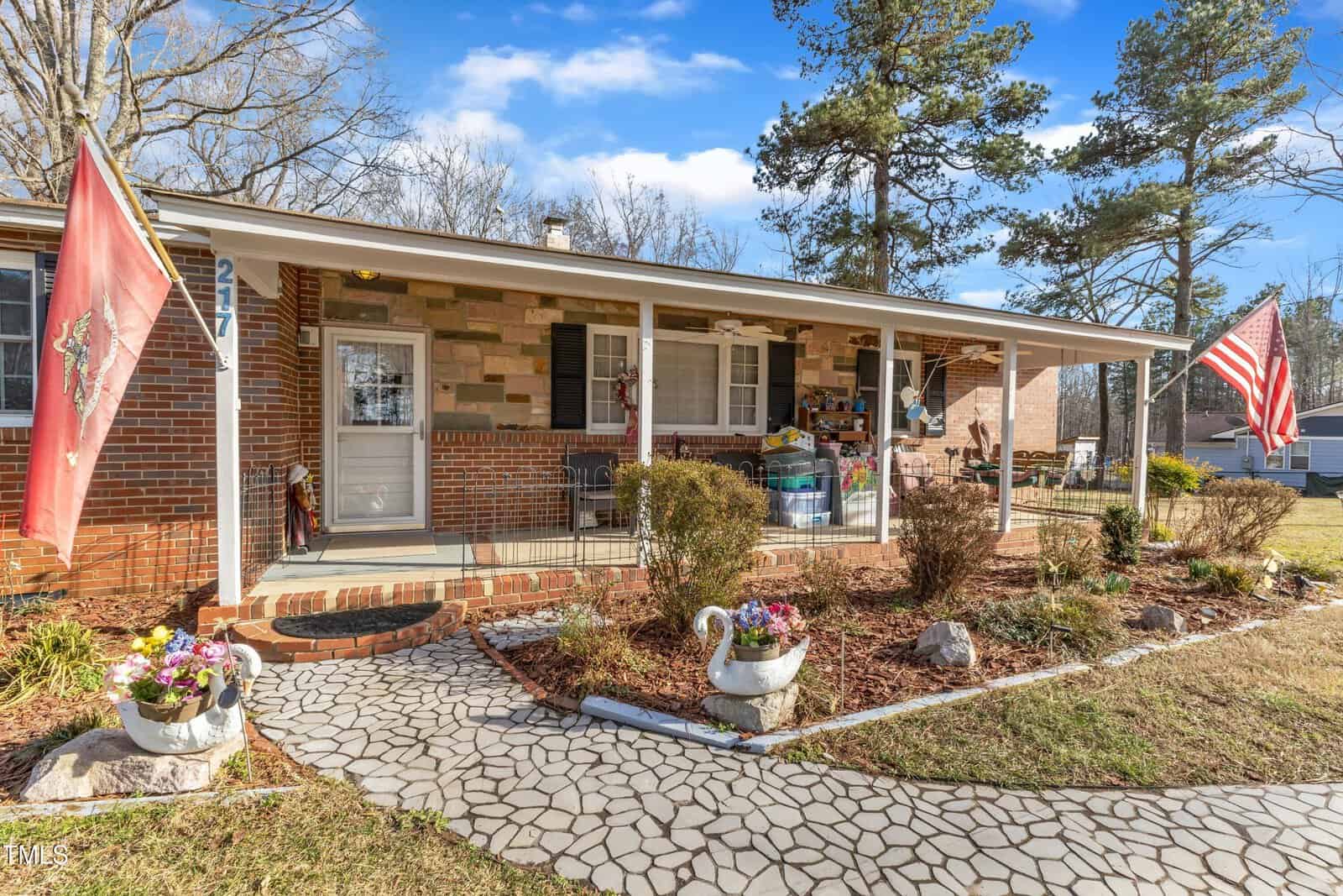 Single-story brick house with a covered front porch, two flags, potted plants, outdoor decorations, and a patterned walkway leading to the entrance. The yard features small shrubs and trees under a blue sky.
