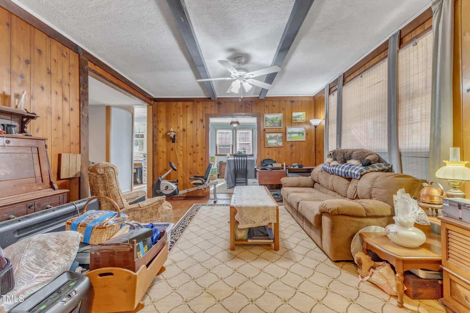 A living room with wood-paneled walls, beige carpet, and a ceiling fan. There is a brown sofa, a plaid blanket, a coffee table, various decorations, and exercise equipment visible in the next room.