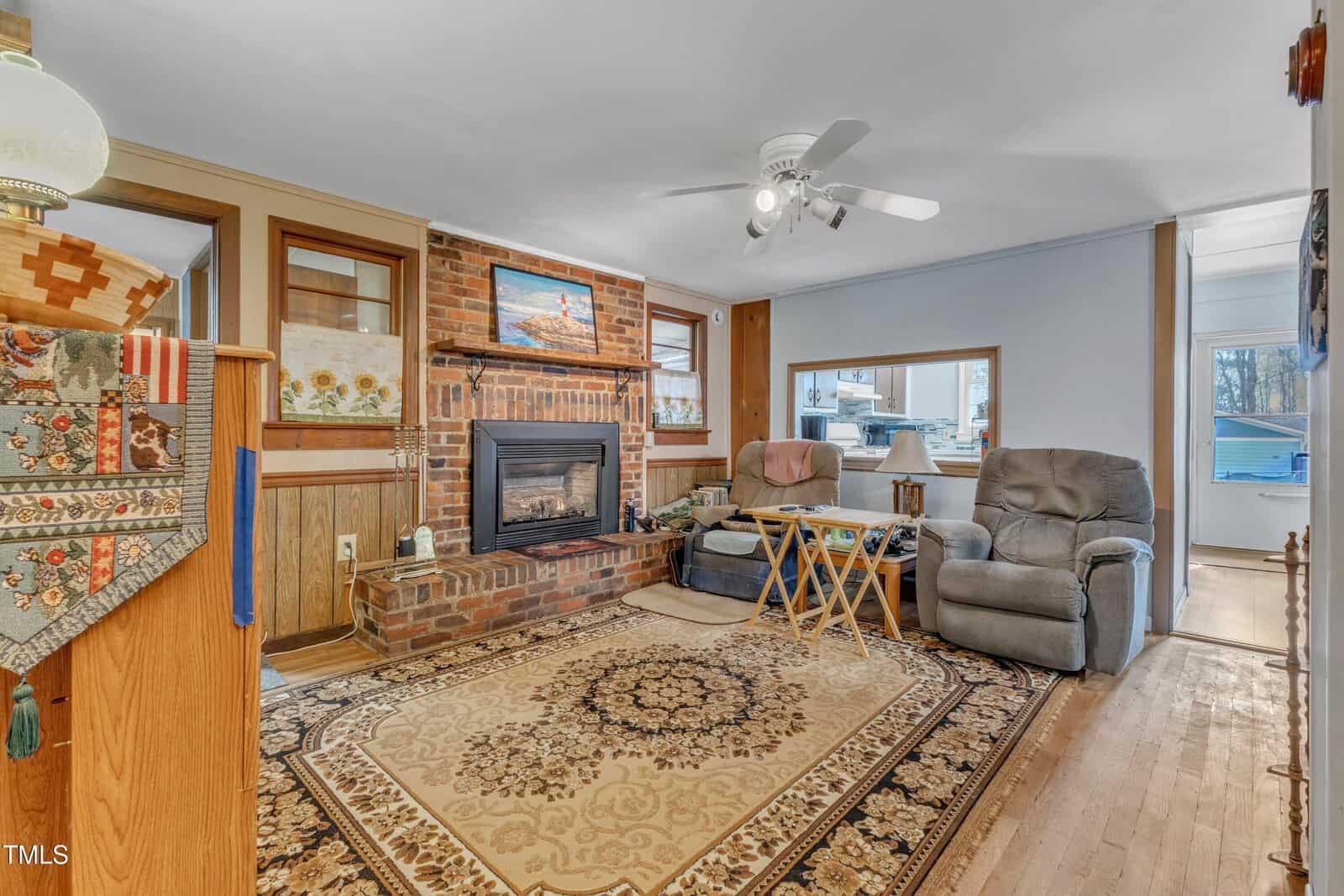 A cozy living room with a brick fireplace, an area rug, two recliners, a small table and tray, wood-paneled walls, hardwood floor, and a ceiling fan. A window opens into another room.