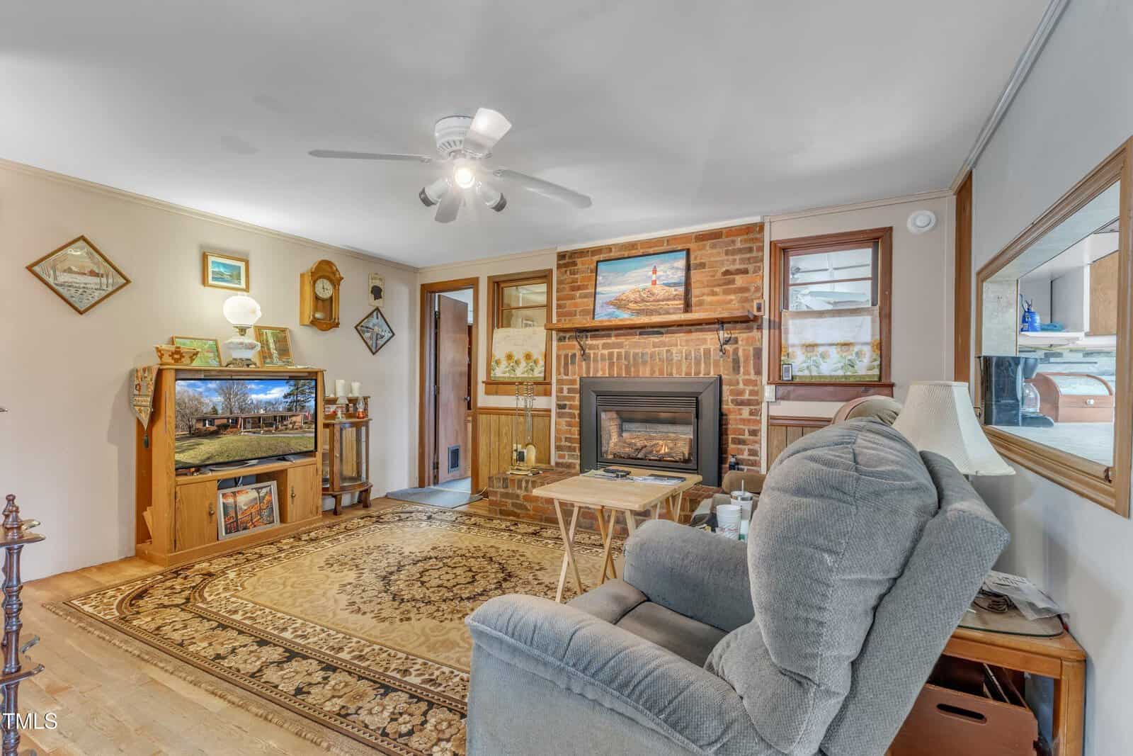 A cozy living room with a patterned rug, armchair, fireplace set in a brick wall, wooden furniture, ceiling fan with lights, wall art, and a TV in a wooden cabinet. A window and open doorway are visible.