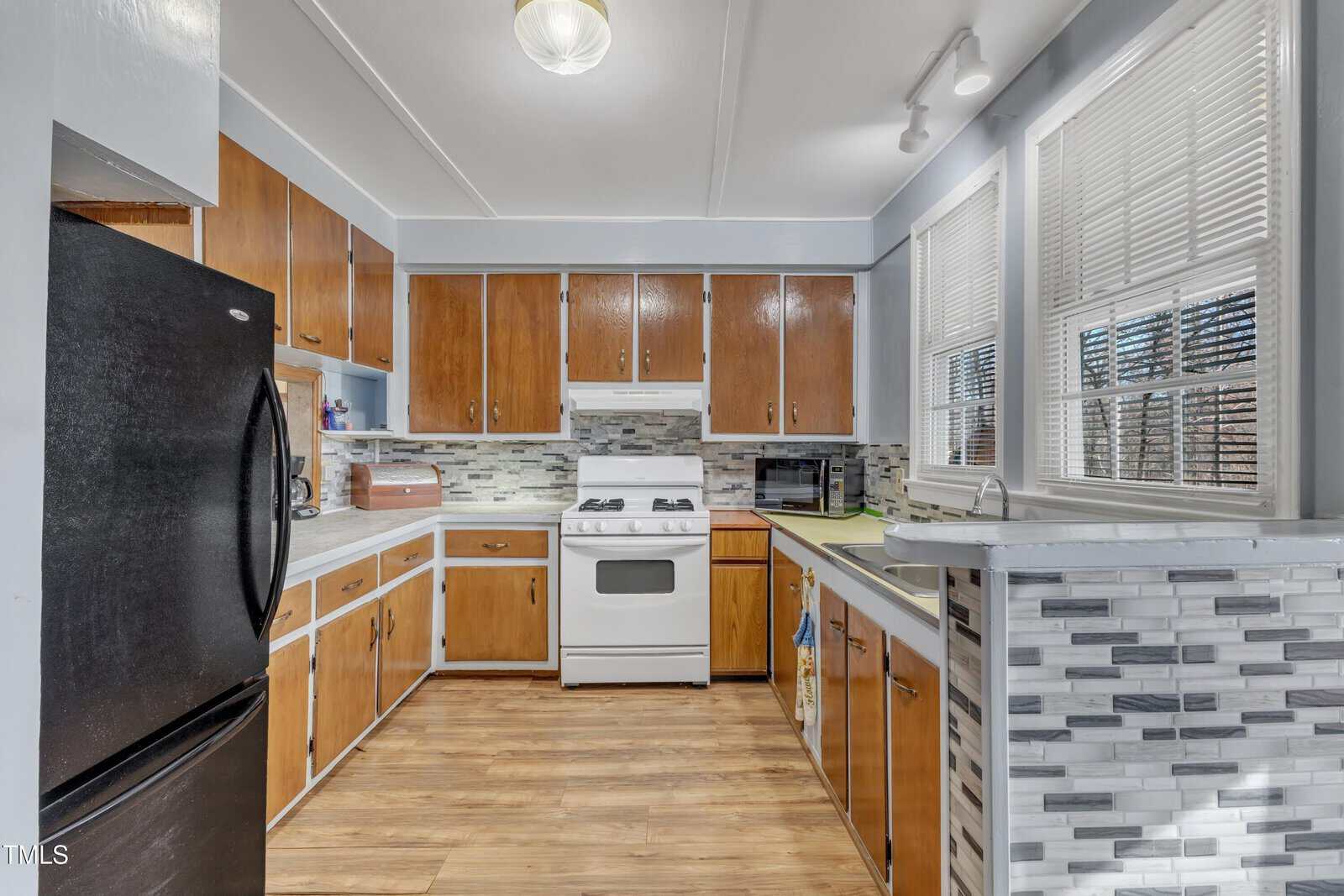 Kitchen with wooden cabinets, white gas stove, black refrigerator, microwave, and toaster on the counter. Gray patterned backsplash and light wood flooring. Two windows let in natural light.