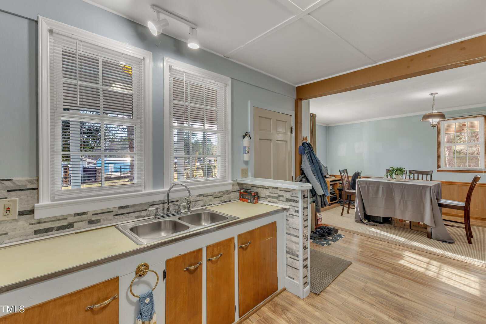 Kitchen with a double sink and wood cabinets, separated from a dining area by a half wall. Light blue walls, windows with blinds, hardwood floors, and a small dining table with chairs are visible.