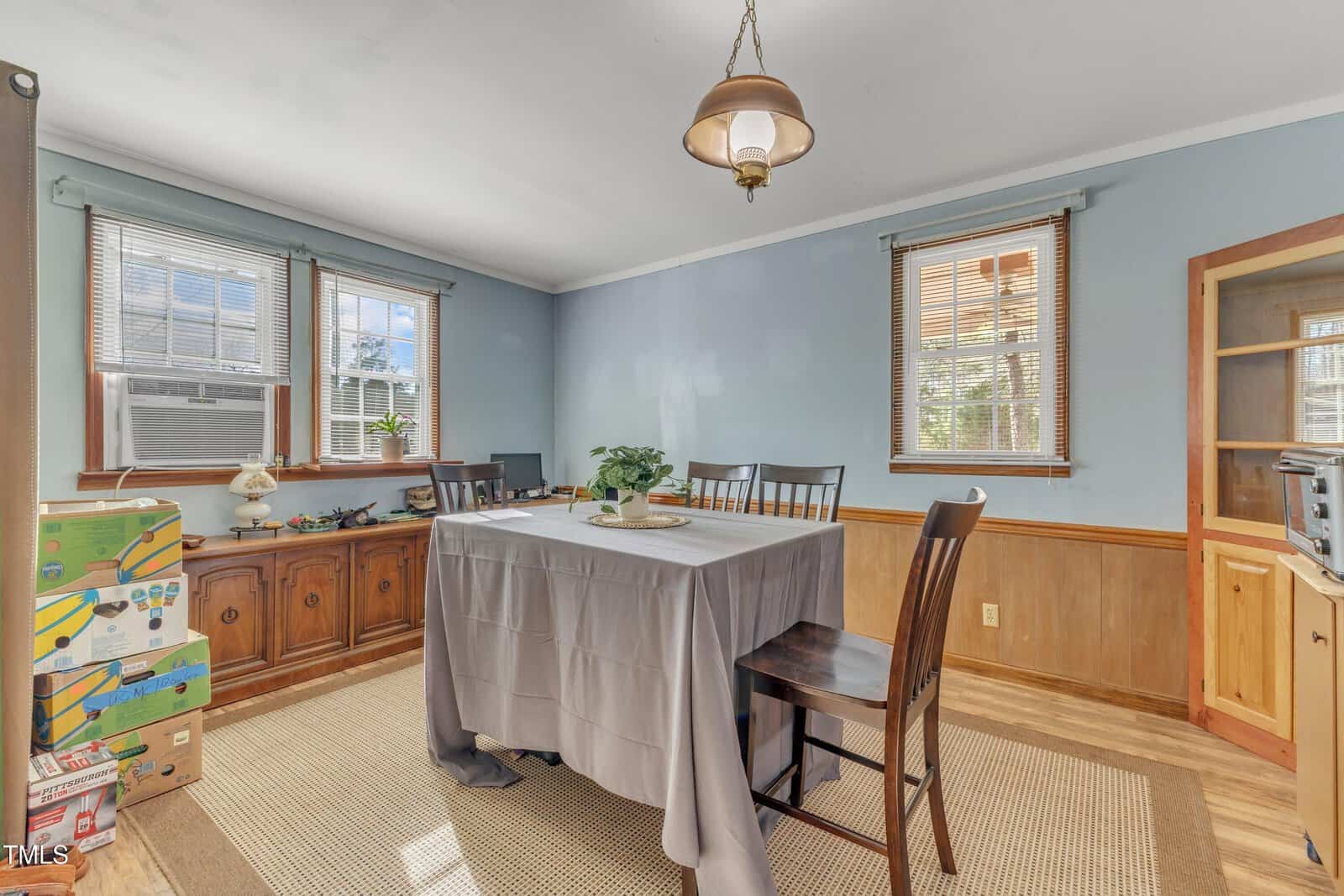 A dining room with blue walls, wood wainscoting, and a table covered by a gray cloth. Four chairs surround the table. Two windows, light fixture, boxes, and sideboard are present. Sunlight enters through the windows.