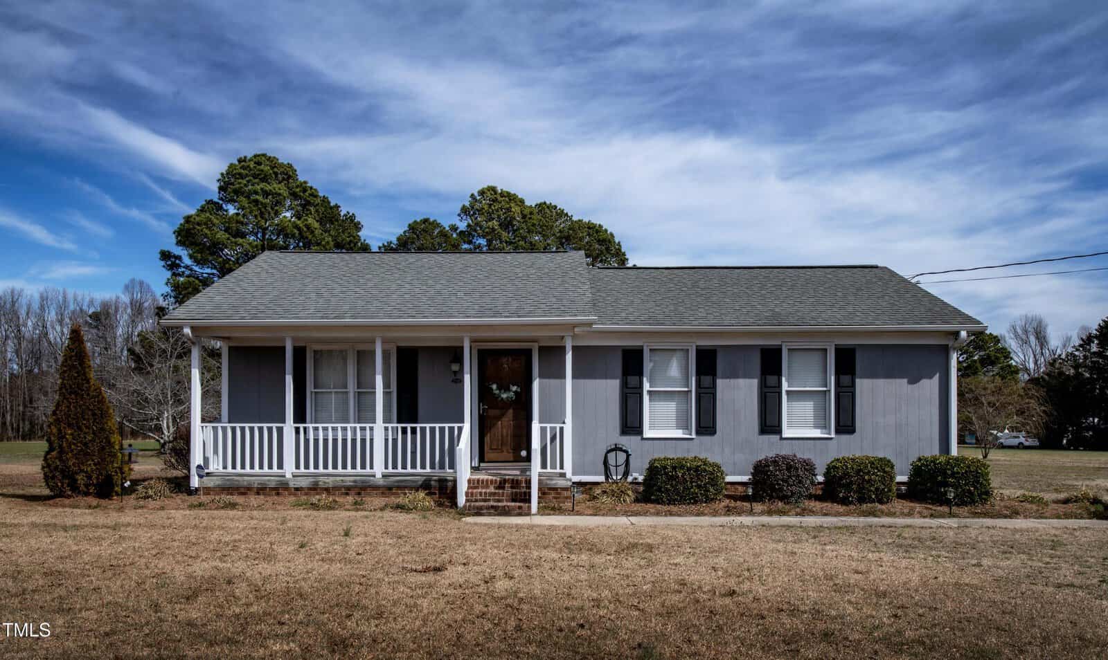 A single-story gray house with white trim and a small front porch is shown. The yard is mostly brown grass with a few shrubs near the house. Pine trees and open sky are visible in the background.