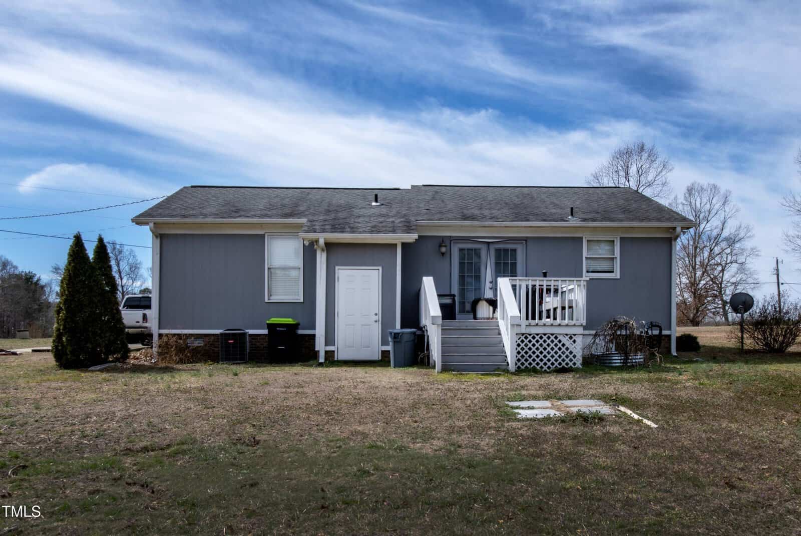 A single-story gray house with white trim, a small back porch with steps, and a white door. The yard is mostly bare with patches of grass, and the sky is partly cloudy.
