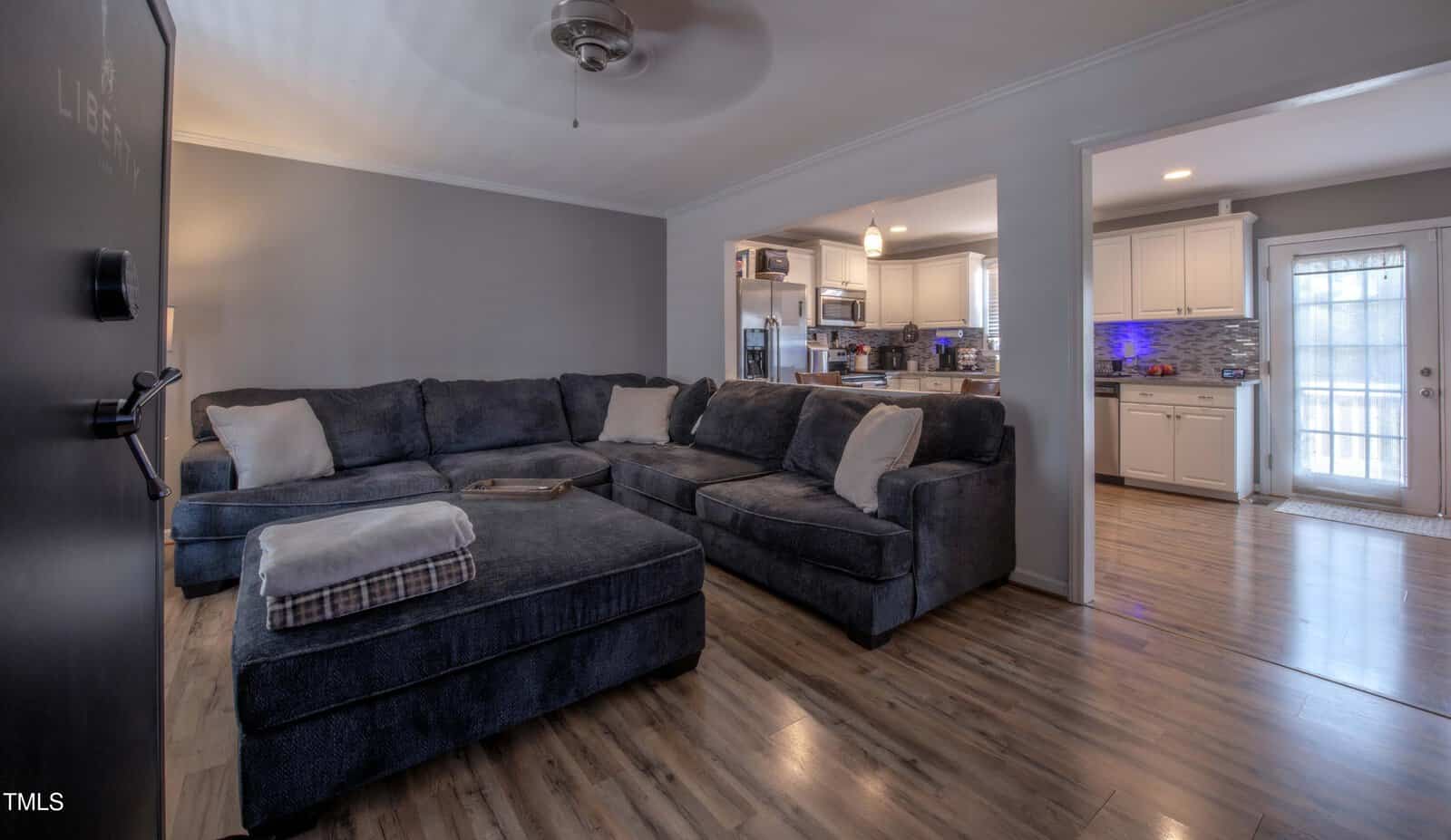 A living room with a large dark gray sectional sofa, wood flooring, and neutral-colored walls. The space opens into a kitchen with white cabinets and stainless steel appliances. Natural light enters through a door and windows.