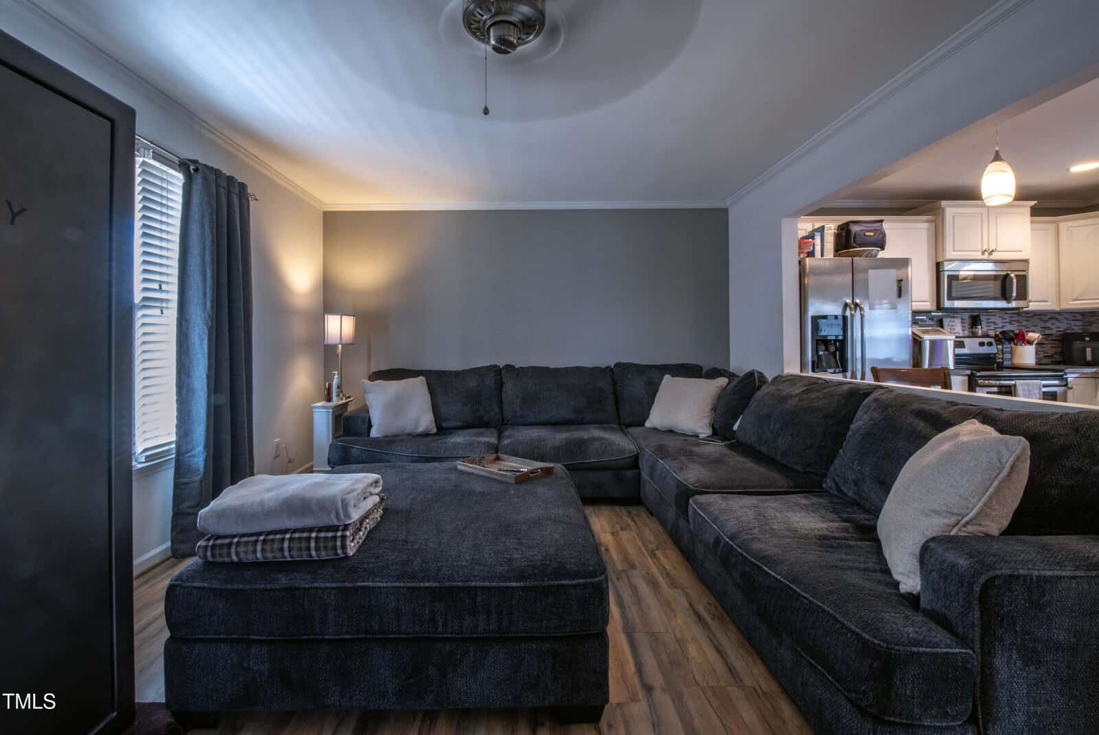 A living room with a large dark gray sectional sofa, a matching ottoman with folded blankets, wood flooring, a standing lamp, and an open view into a kitchen with white cabinets and stainless steel appliances.