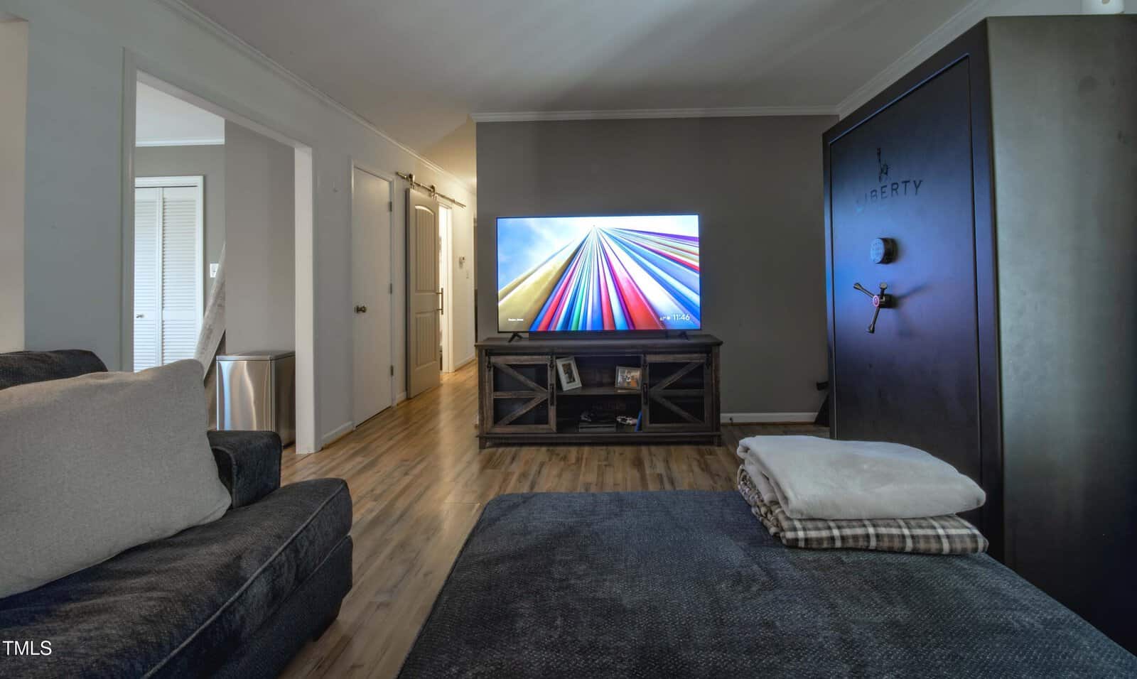 A living room with a dark sofa, folded blankets on a table, a large flat-screen TV displaying colorful lines, a wooden TV stand, and a tall dark safe labeled "Liberty" against a gray wall. Hardwood floors throughout.