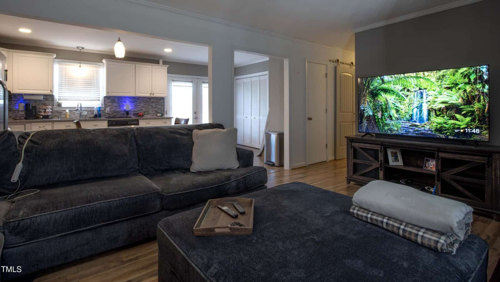 A living room with a dark gray sectional sofa, ottoman, and a tray with remotes. A TV on a wooden stand shows a nature scene. Kitchen with white cabinets is visible in the background. Hardwood floors throughout.