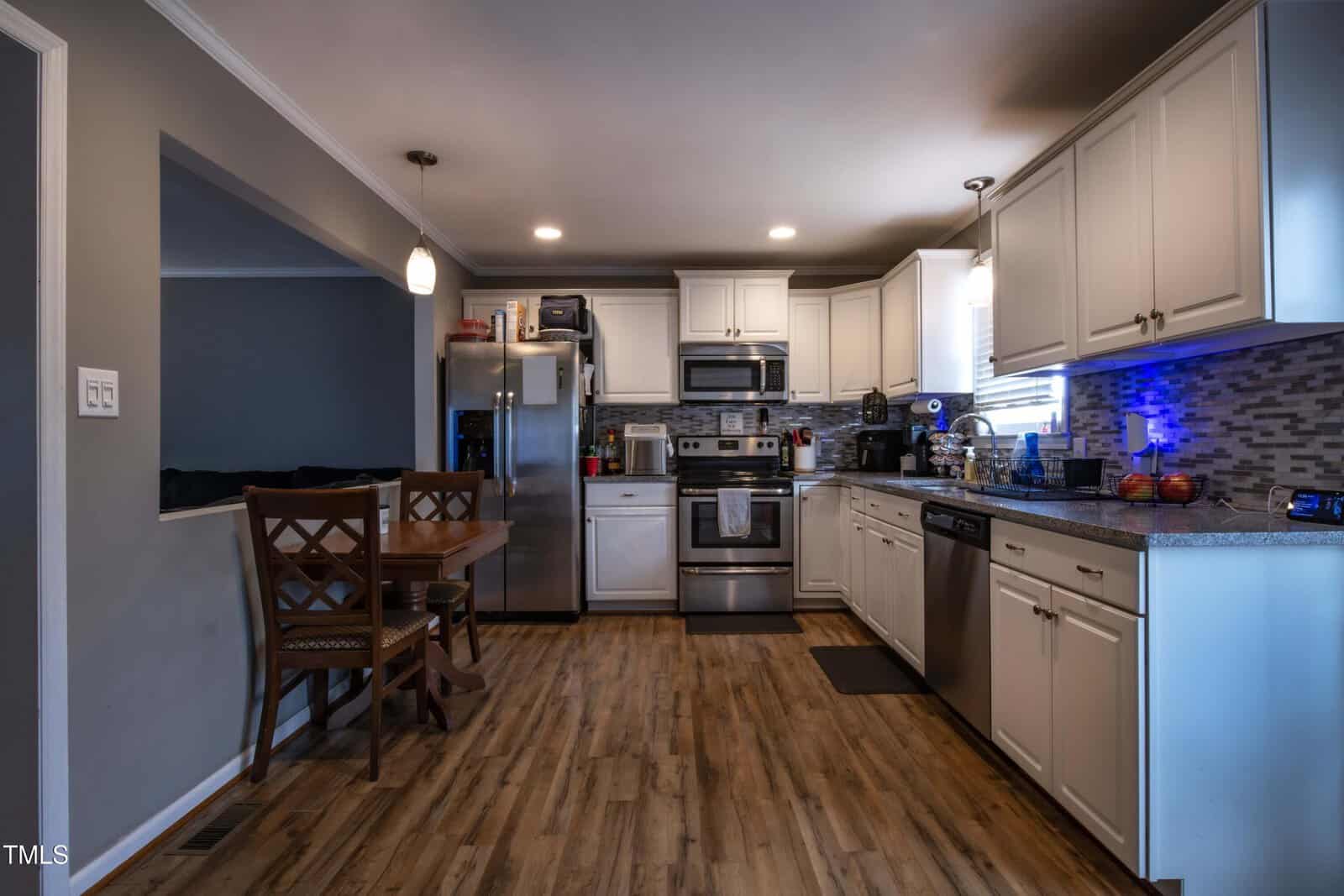 Modern kitchen with white cabinets, stainless steel appliances, wood-style flooring, a small dining table with four chairs, and tile backsplash. Countertops hold various kitchen items. Room is well-lit with ceiling and pendant lights.
