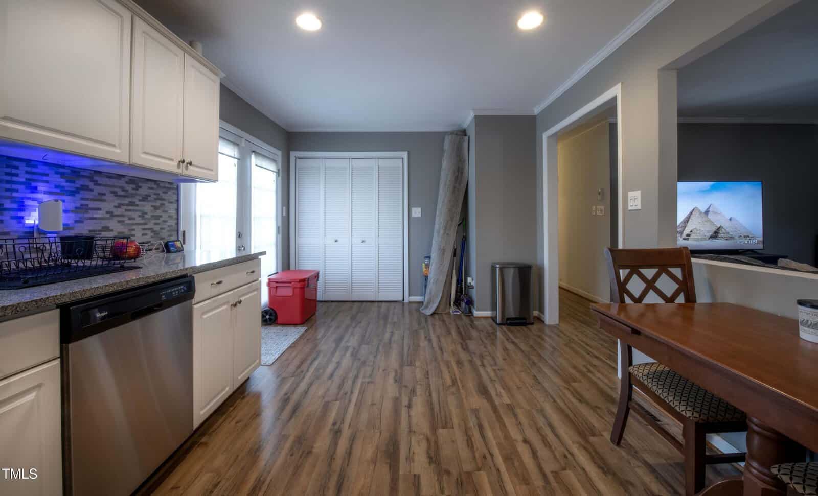 A kitchen with white cabinets, a dishwasher, and tile backsplash opens to a dining area with a wooden table. Sliding doors and a closet are at the back, with a red storage bin and cleaning items visible. A TV is on in the next room.
