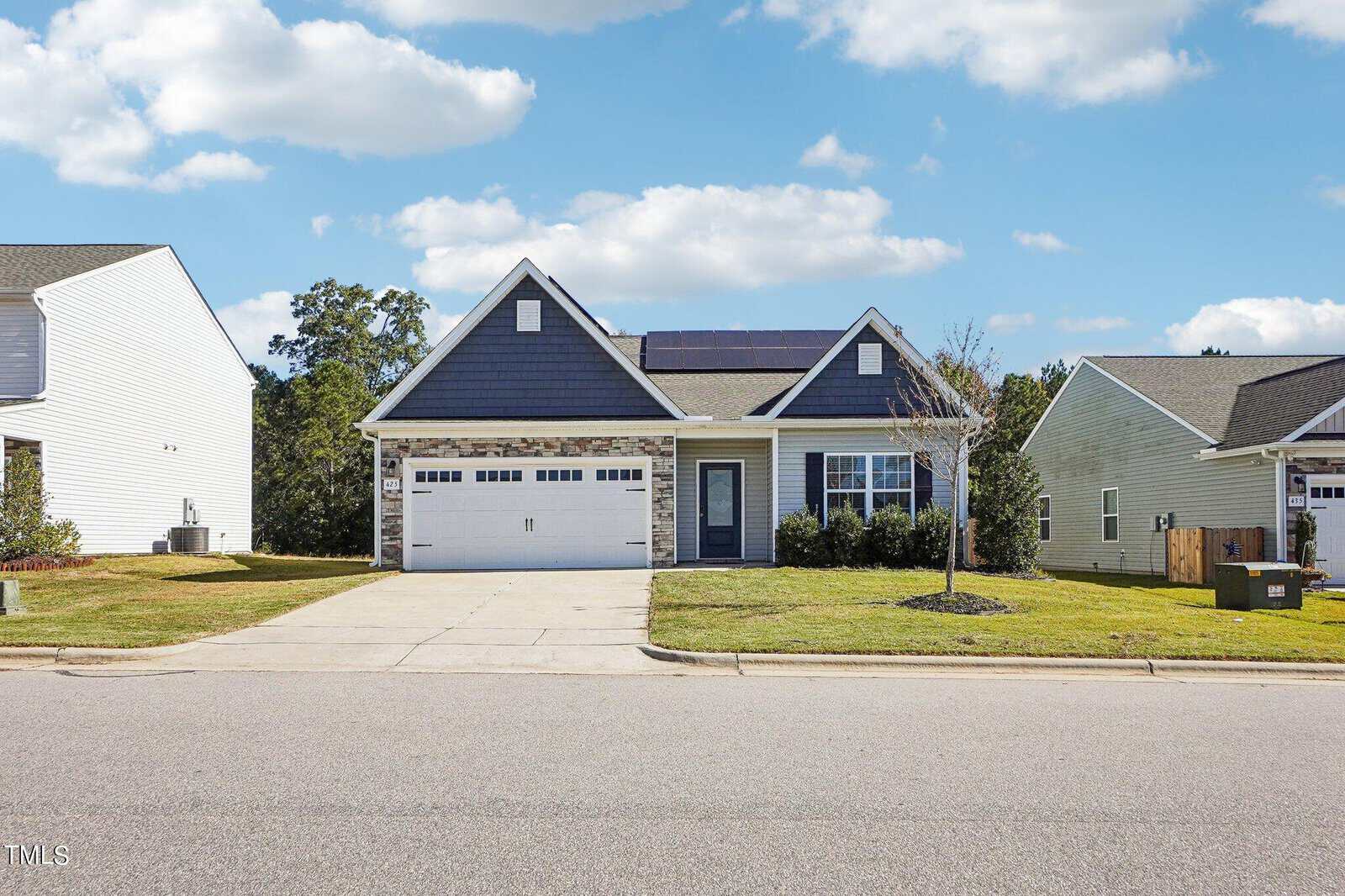 A single-story house with blue siding, stone accents around the garage, solar panels on the roof, a double garage door, a small front porch, and a neatly trimmed lawn, set in a suburban neighborhood under a partly cloudy sky.