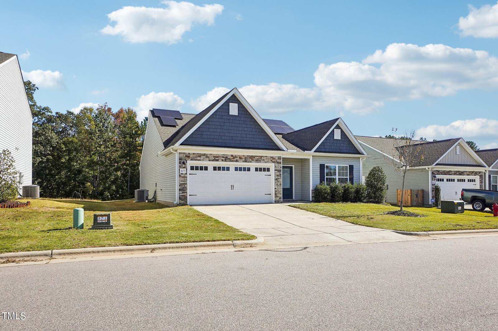 Single-story suburban house with a double garage, light-colored siding, dark roof shingles, and stone accents above the garage. The property has a driveway, front lawn, and is situated on a quiet street with similar houses nearby.