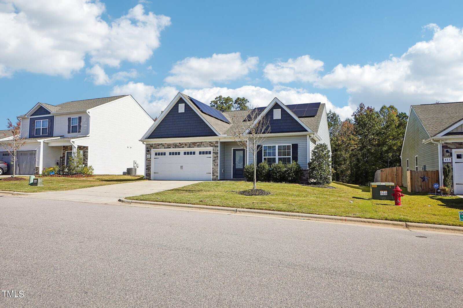A single-story house with blue siding, white trim, and a two-car garage sits on a lawn next to similar houses. There are solar panels on the roof and a fire hydrant near the curb. The sky is partly cloudy.