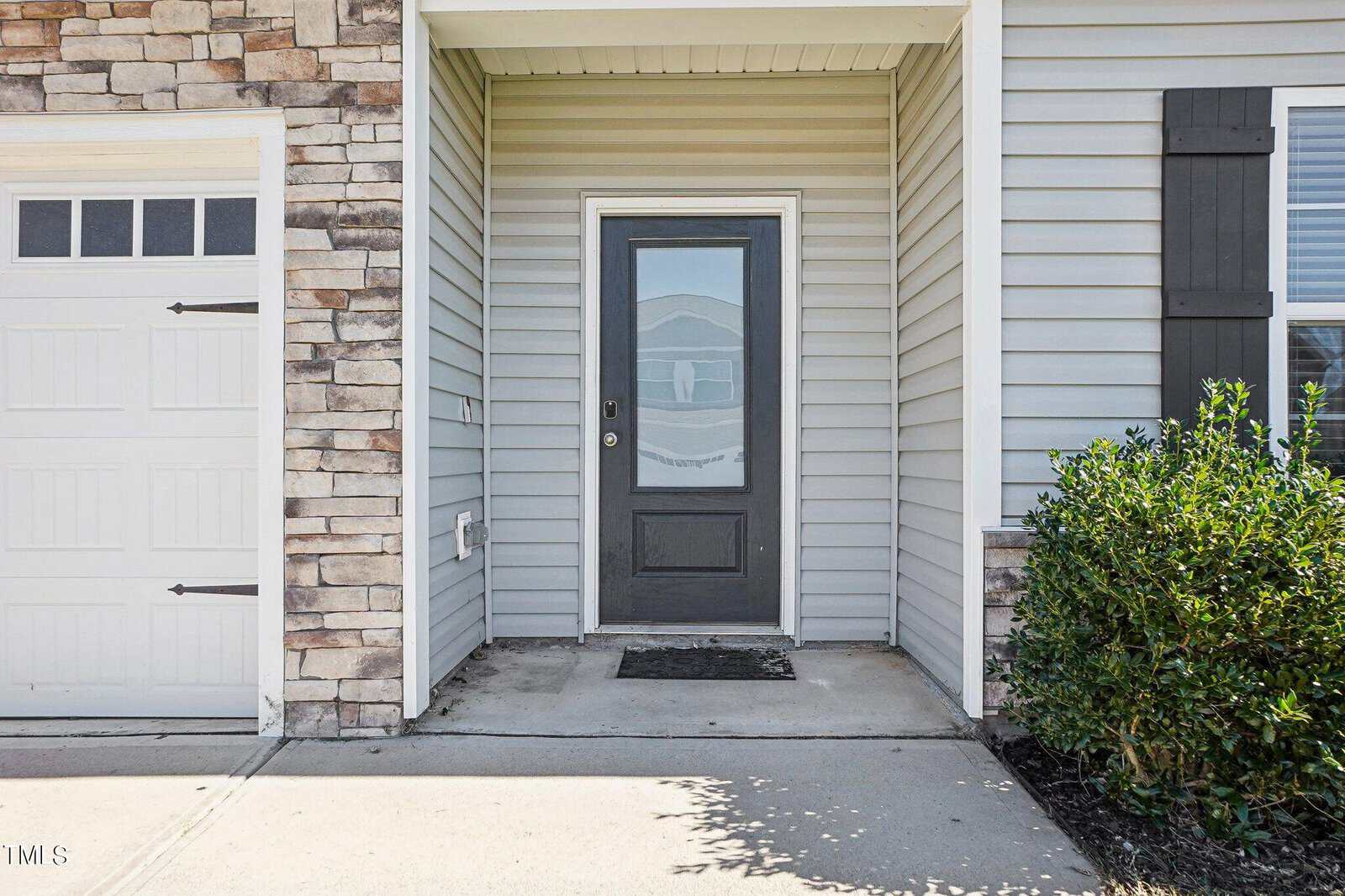 A gray front door with a glass panel is centered between beige siding and a stone wall. A garage door is on the left, and green bushes are on the right. A doormat sits in front of the entryway.
