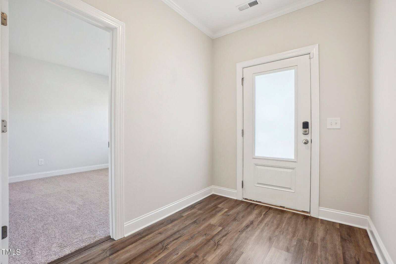 A small entryway with light-colored walls, wood-look flooring, and a white door with a frosted glass panel. An adjacent carpeted room is visible through an open doorway on the left.