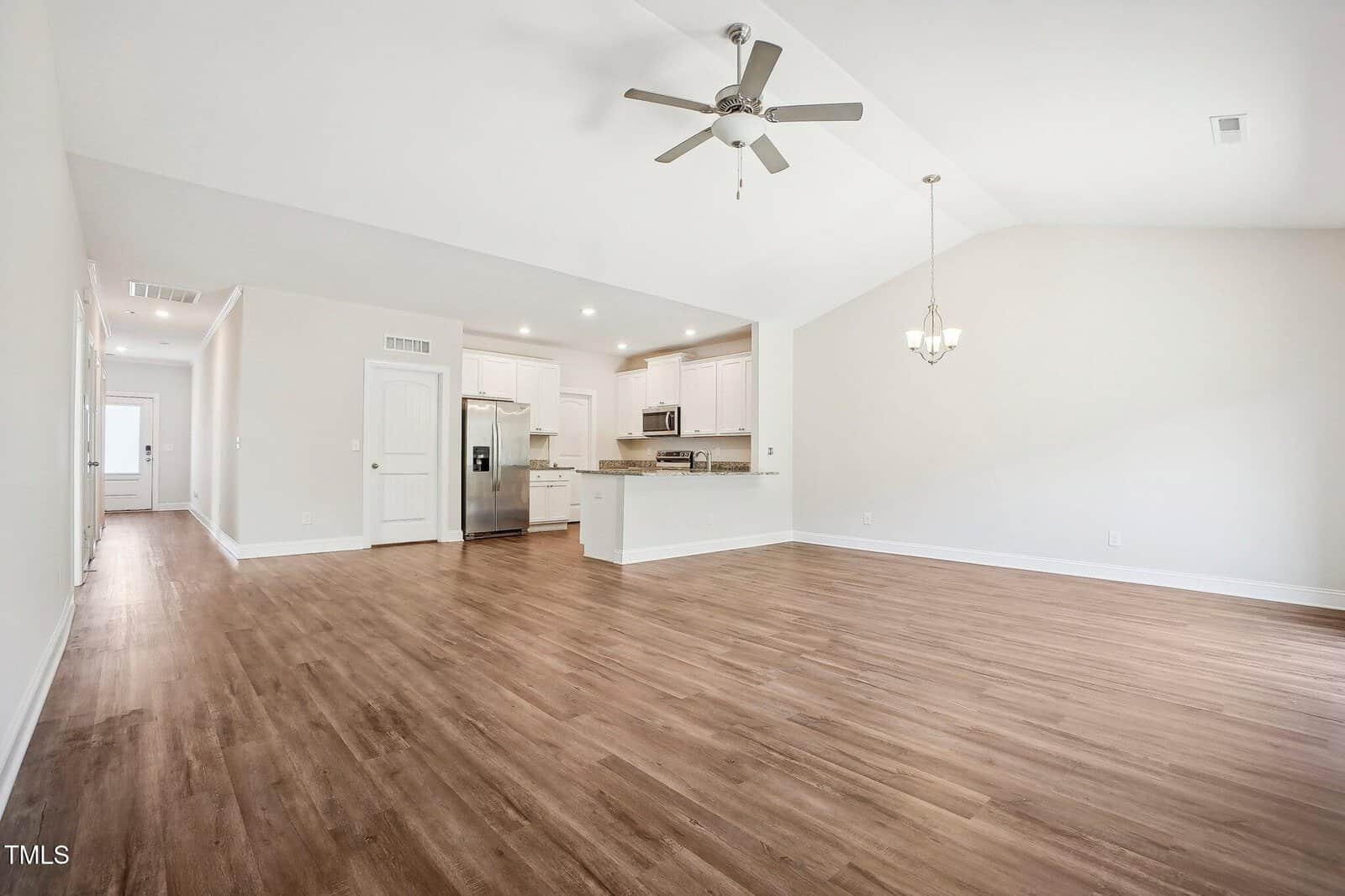 Spacious open-concept living area with light wood flooring, white walls, a ceiling fan, and a chandelier. The kitchen features white cabinets, stainless steel appliances, and recessed lighting. A hallway is visible in the background.