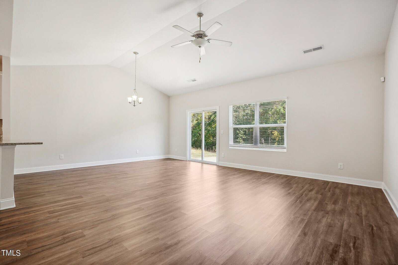 A spacious, empty living room with light wood flooring, white walls, a ceiling fan, a chandelier, two windows, and a glass door leading outside.