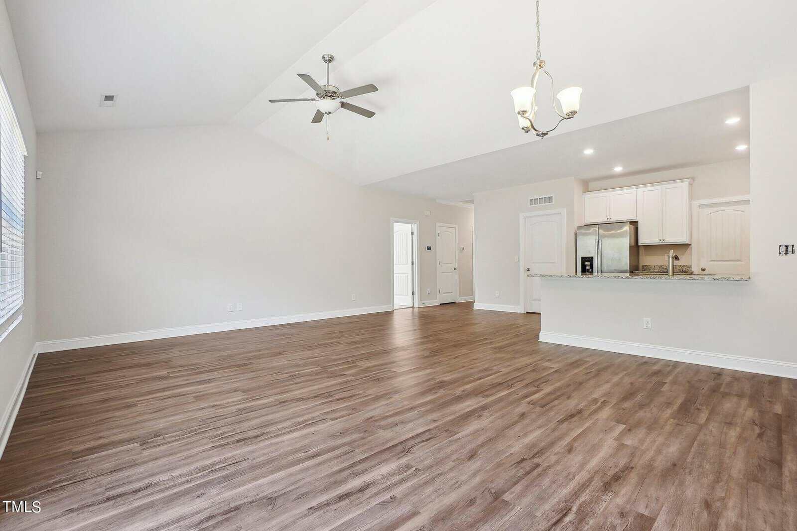 Spacious empty living area with wood flooring, white walls, ceiling fan, chandelier, and an open kitchen featuring white cabinets and stainless steel appliances. Large windows provide natural light.