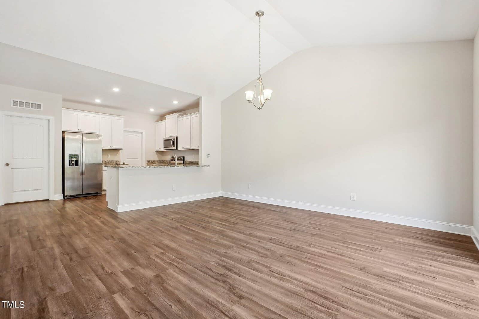 Unfurnished open-plan room with light-colored walls and wood flooring, featuring a kitchen area with white cabinets, stainless steel appliances, and a breakfast bar. A chandelier hangs over the dining space.