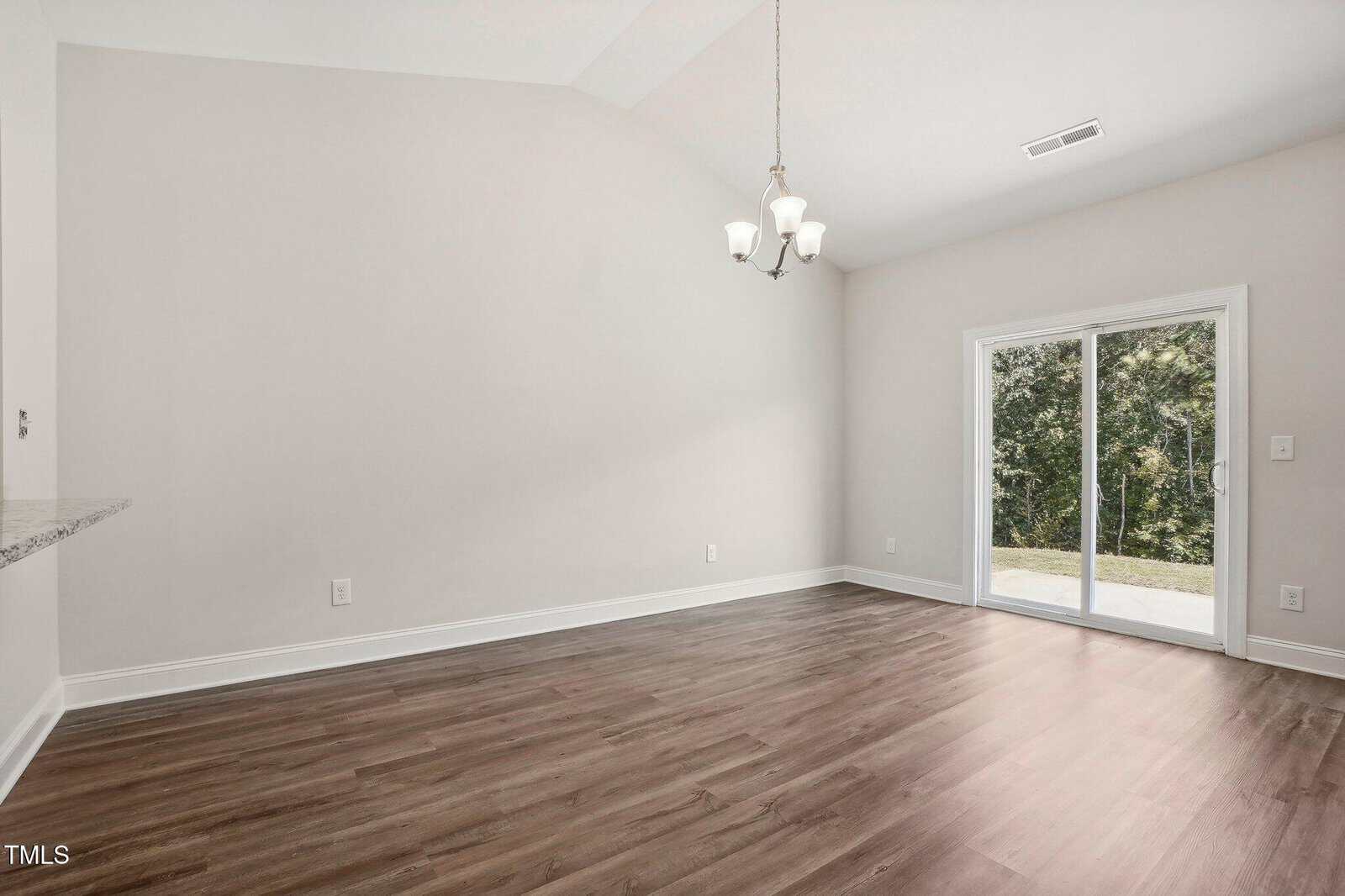 A bright empty room with light-colored walls, wood flooring, a ceiling light fixture, and sliding glass doors leading to an outdoor area with trees visible outside.