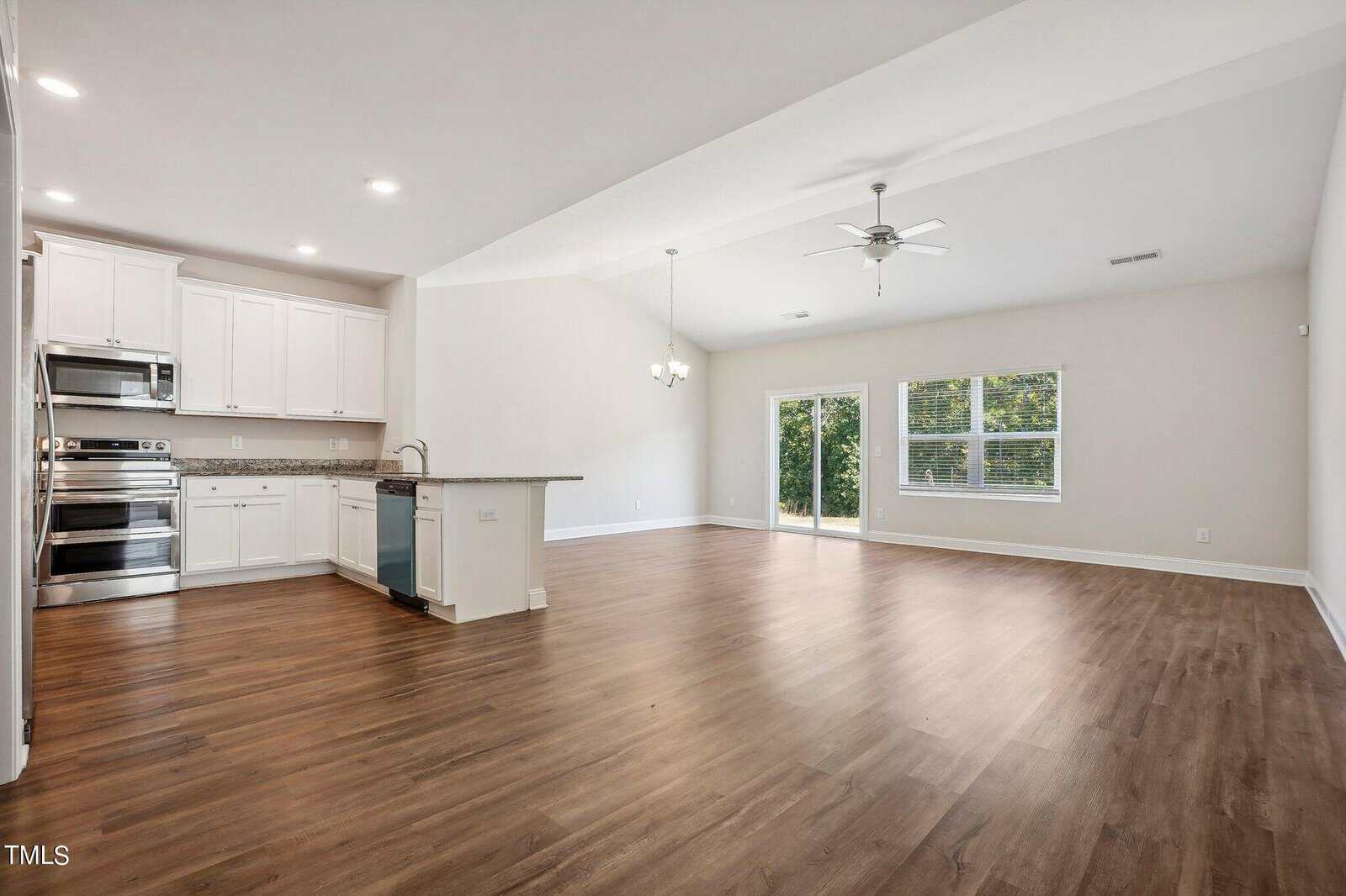 Spacious open-concept living area with wood floors, white kitchen cabinets, stainless steel appliances, granite countertops, and large windows overlooking greenery, plus a ceiling fan and patio doors.