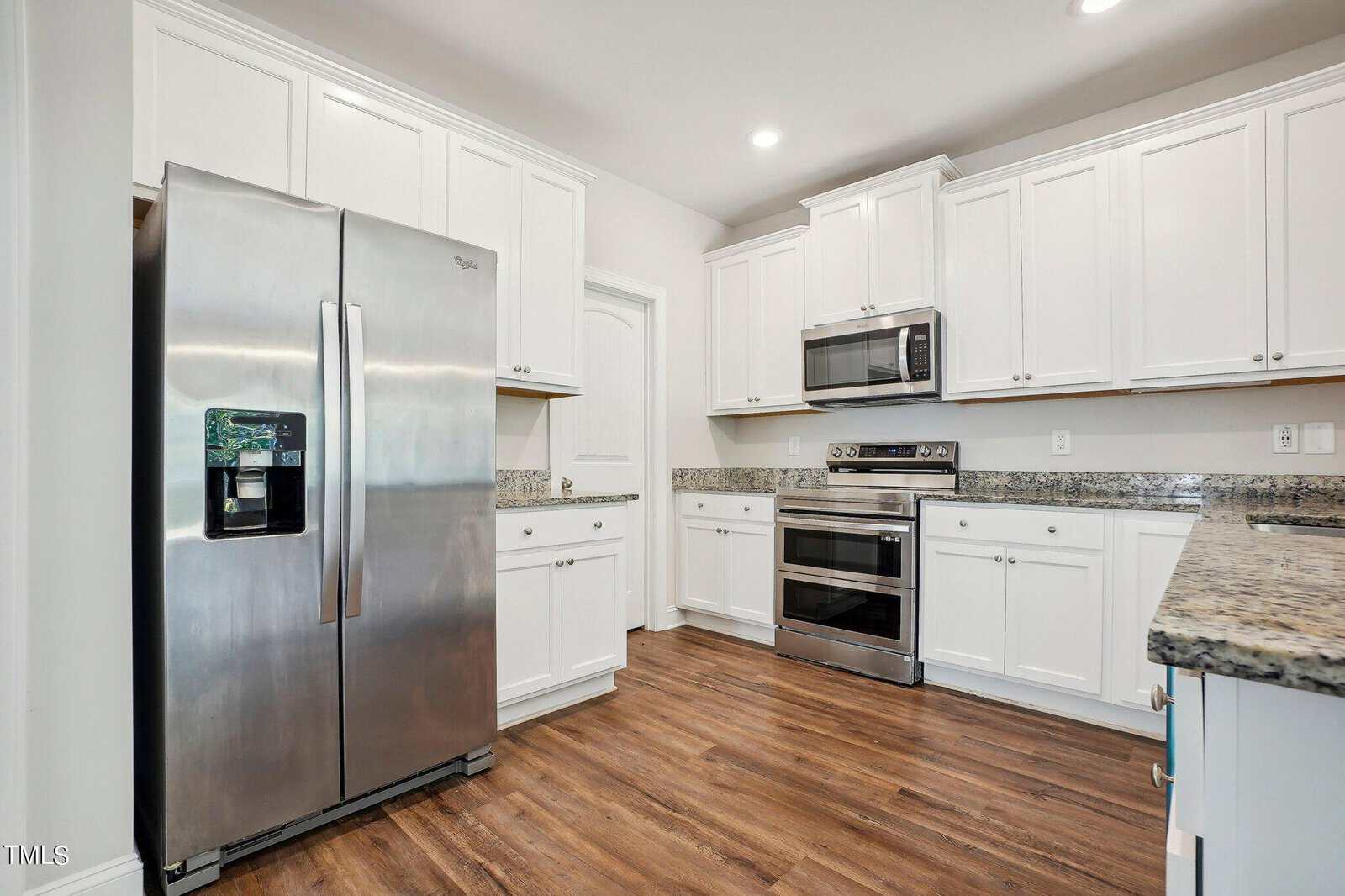 Modern kitchen with white cabinets, stainless steel appliances including a double oven and fridge, granite countertops, and wood-look flooring under bright lighting.