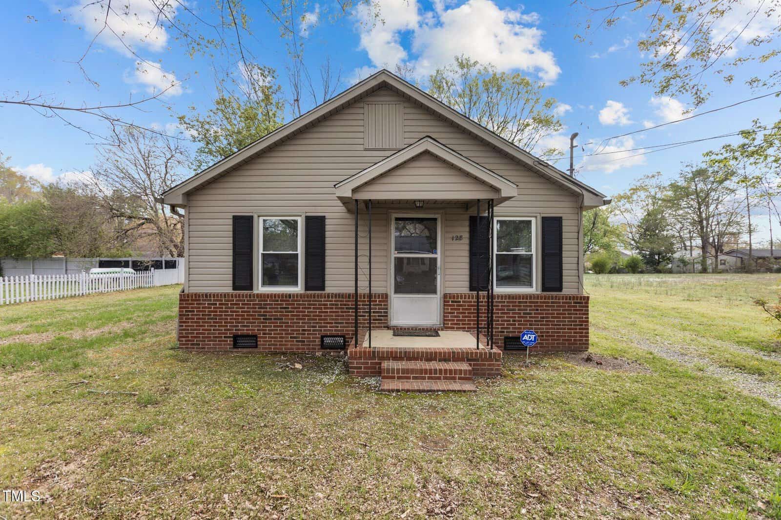 A small beige house with a gable roof at 128 Warren Avenue, Oxford. It features black shutters, a brick foundation, and a covered front porch with steps. The grassy yard has scattered trees, a white picket fence on the left, and a blue ADT security sign by the steps.