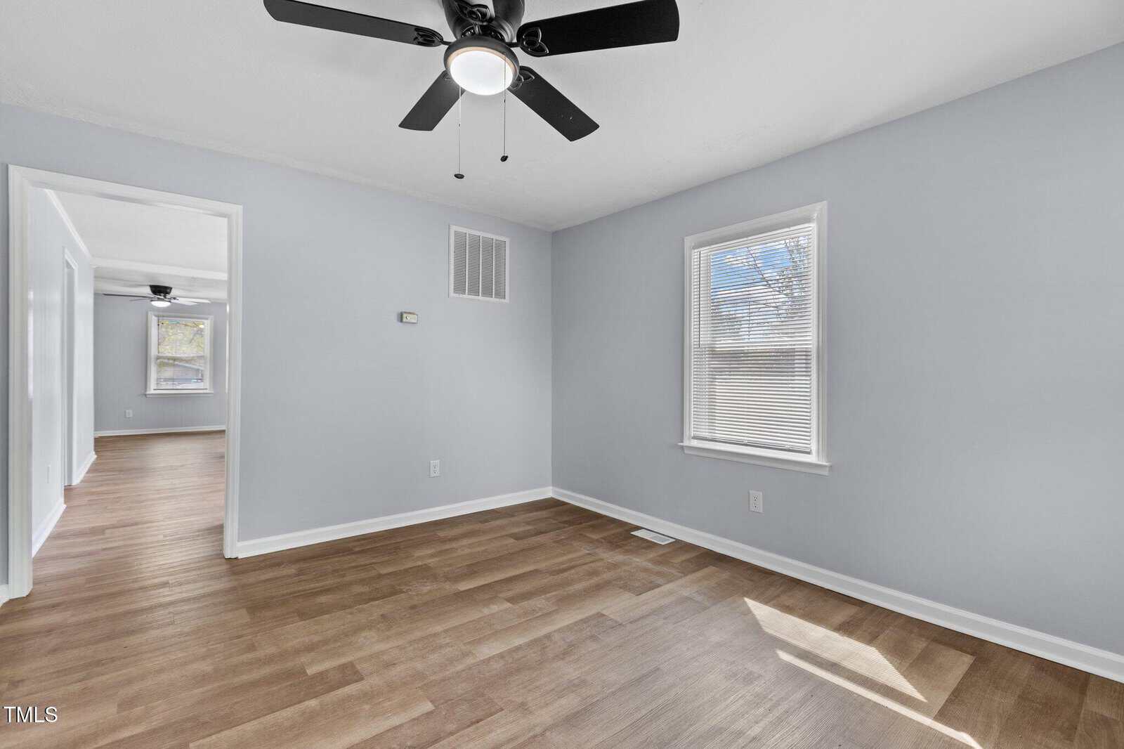 A bright, empty room with light gray walls, wooden floors, a ceiling fan with lights, and a window letting in sunlight. An open doorway leads to another room with similar flooring and wall color.