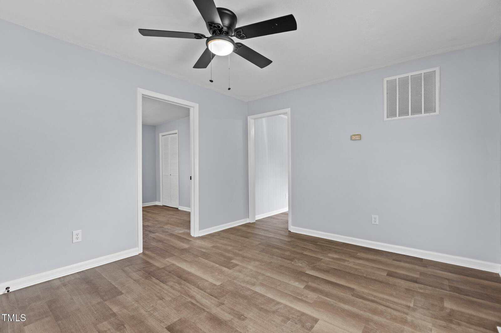 A clean, empty room with light gray walls, wood-style flooring, a black ceiling fan with a light, and two open doorways leading to other areas. White baseboards and vents are visible on the walls.
