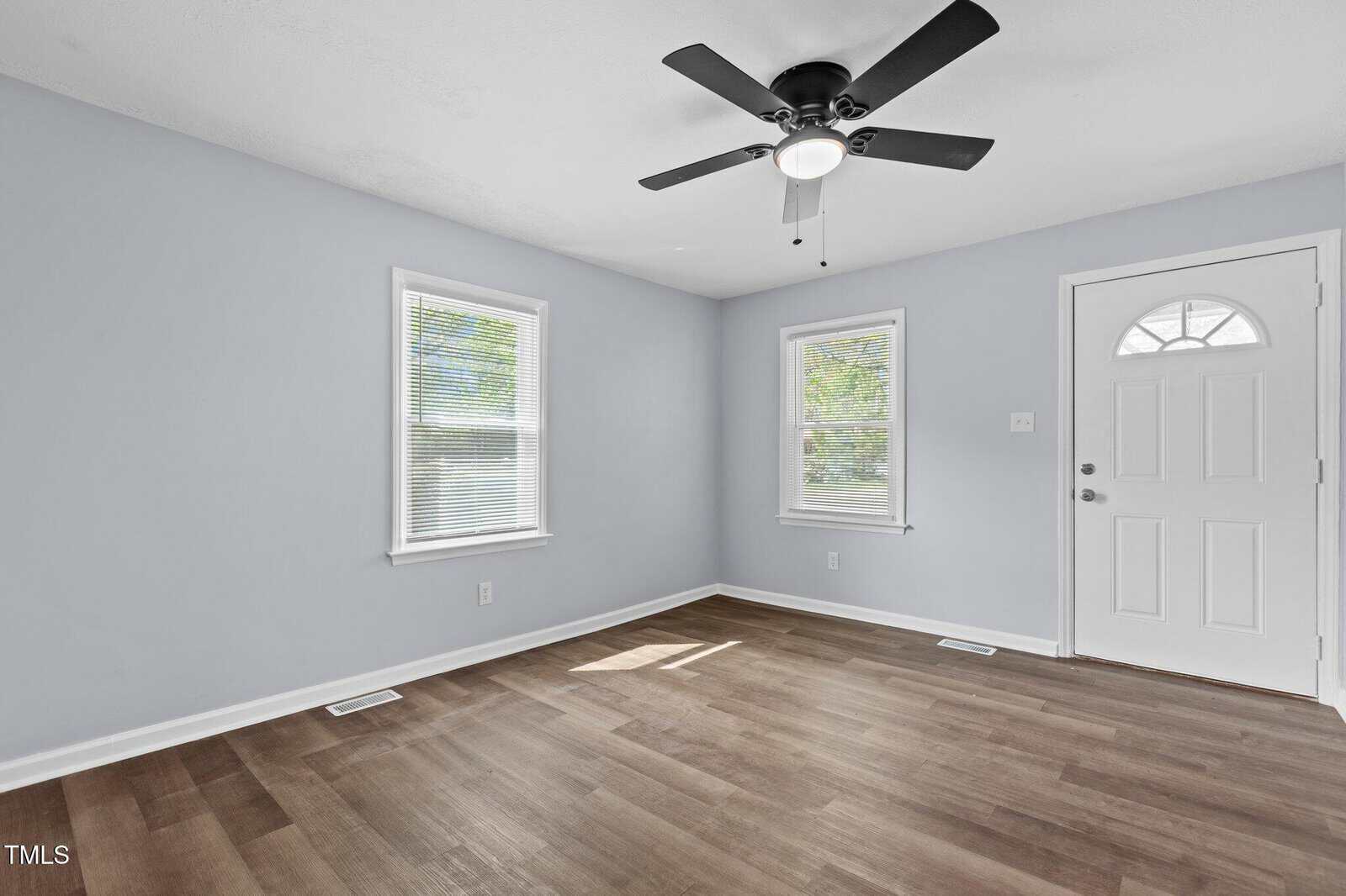A bright, empty room with light gray walls, two windows with blinds, a white door, a black ceiling fan, and wood-look vinyl flooring. Natural light fills the space.