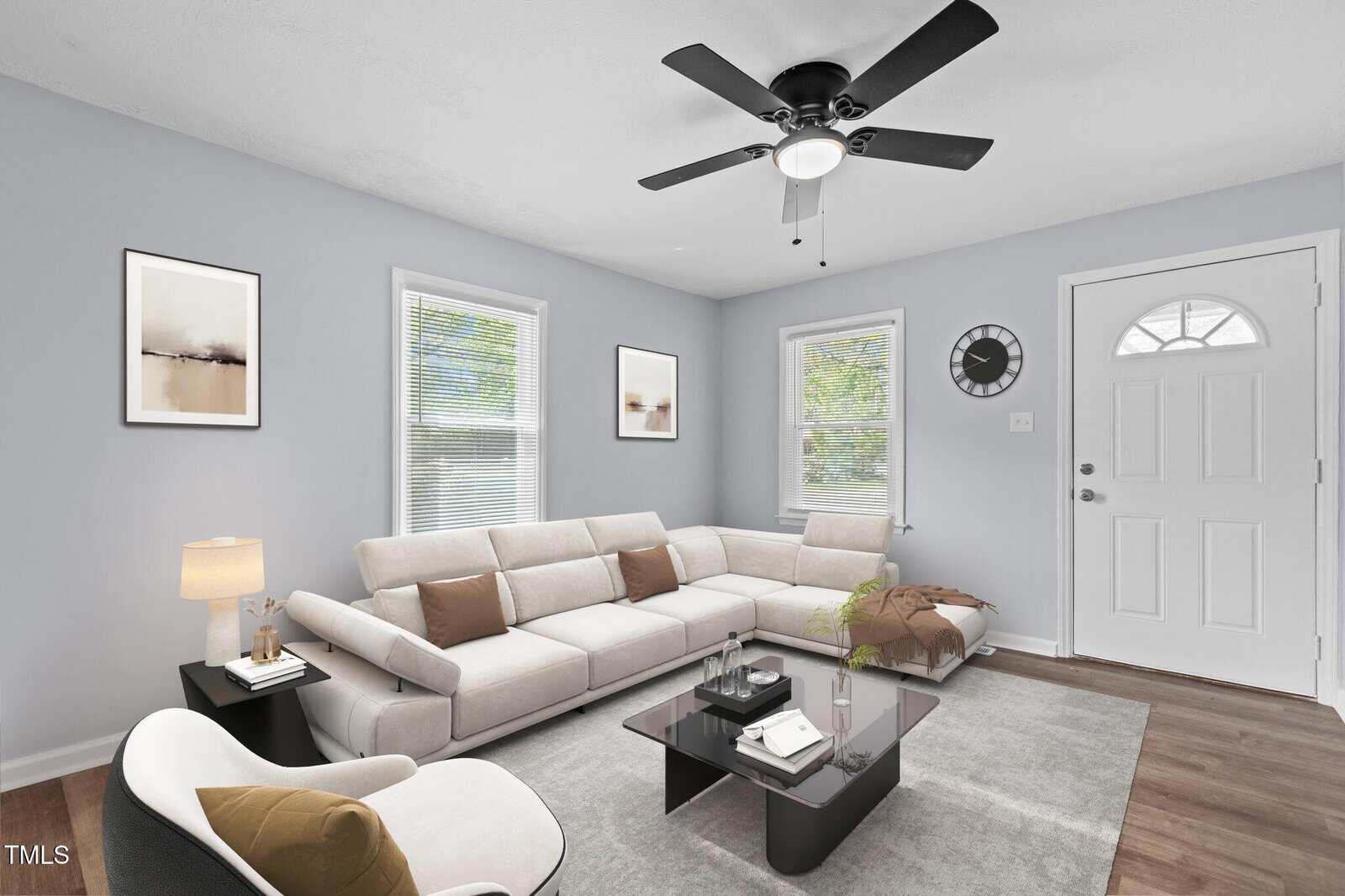 A modern living room with a white sectional sofa, a white armchair, a glass coffee table, wall art, a clock, and two windows. The walls are light gray, and the floor is wood. A ceiling fan is centered above.